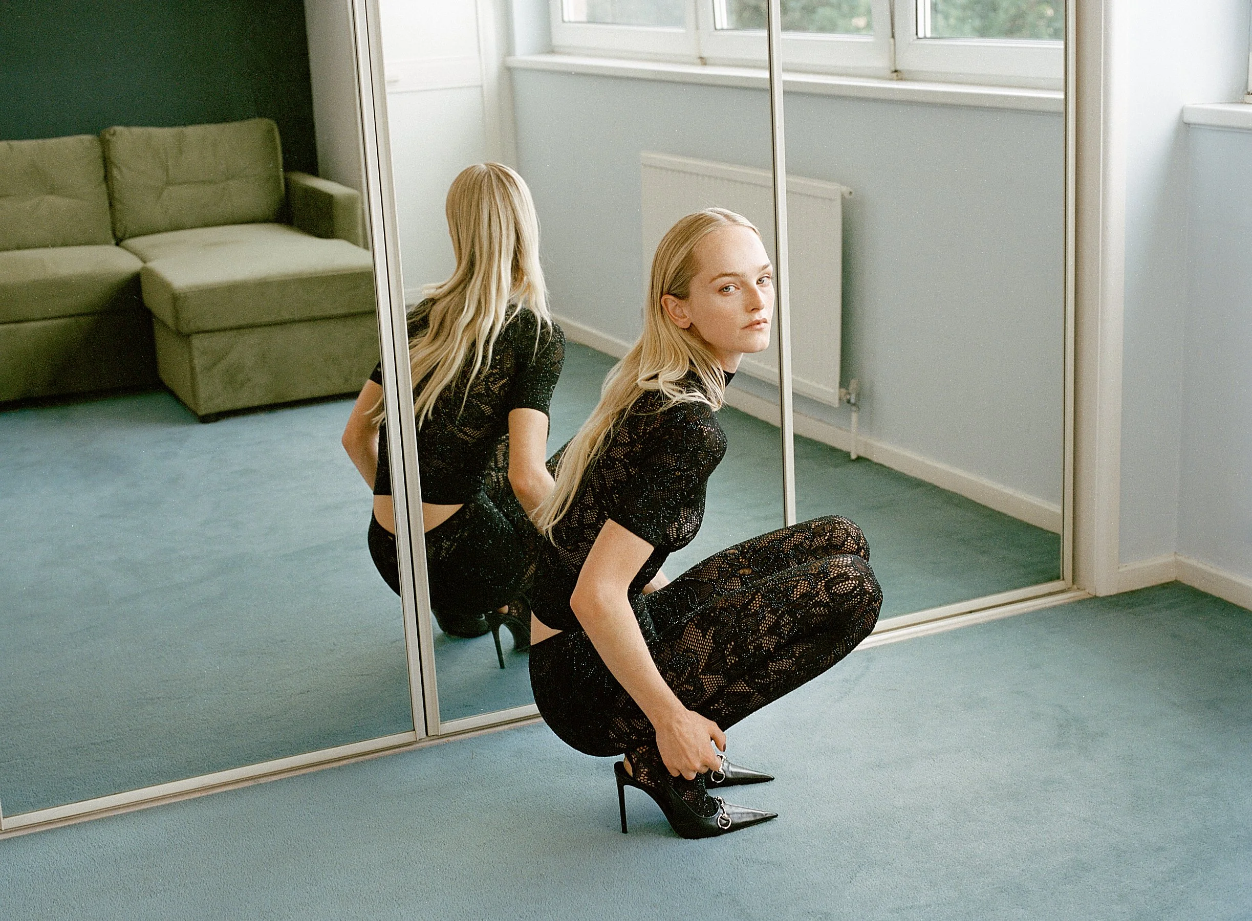 A woman with long blonde hair sits on the floor in front of a mirror, adjusting her black high-heeled shoe. She is wearing a black lace dress and appears to be in a room with a green sofa, a window, and light-colored walls.