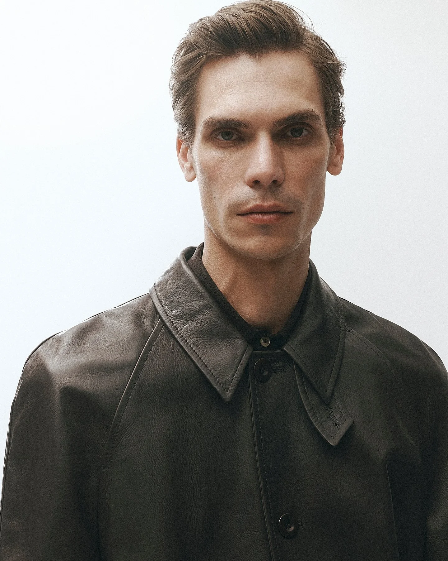 A young man with short, styled brown hair and light skin wearing a black leather jacket against a plain light background.