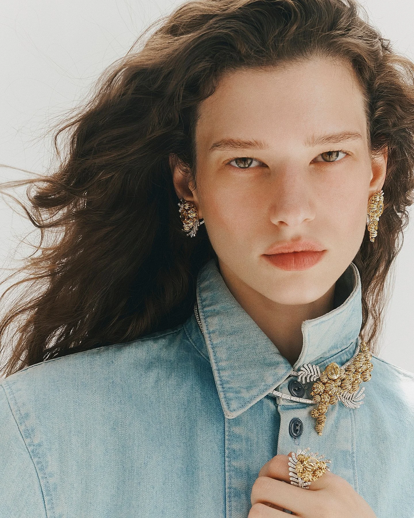A woman with wavy brown hair wearing large gold earrings, a chunky gold necklace, and a gold ring. She is dressed in a light denim shirt and has a neutral background.