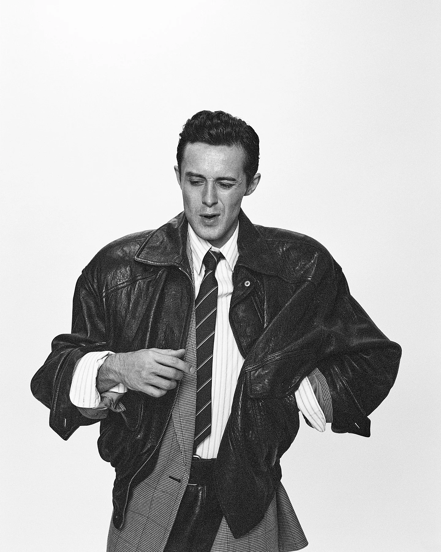 A man in a leather jacket over a striped shirt and tie, with a hat hanging from his belt, standing against a plain white background.