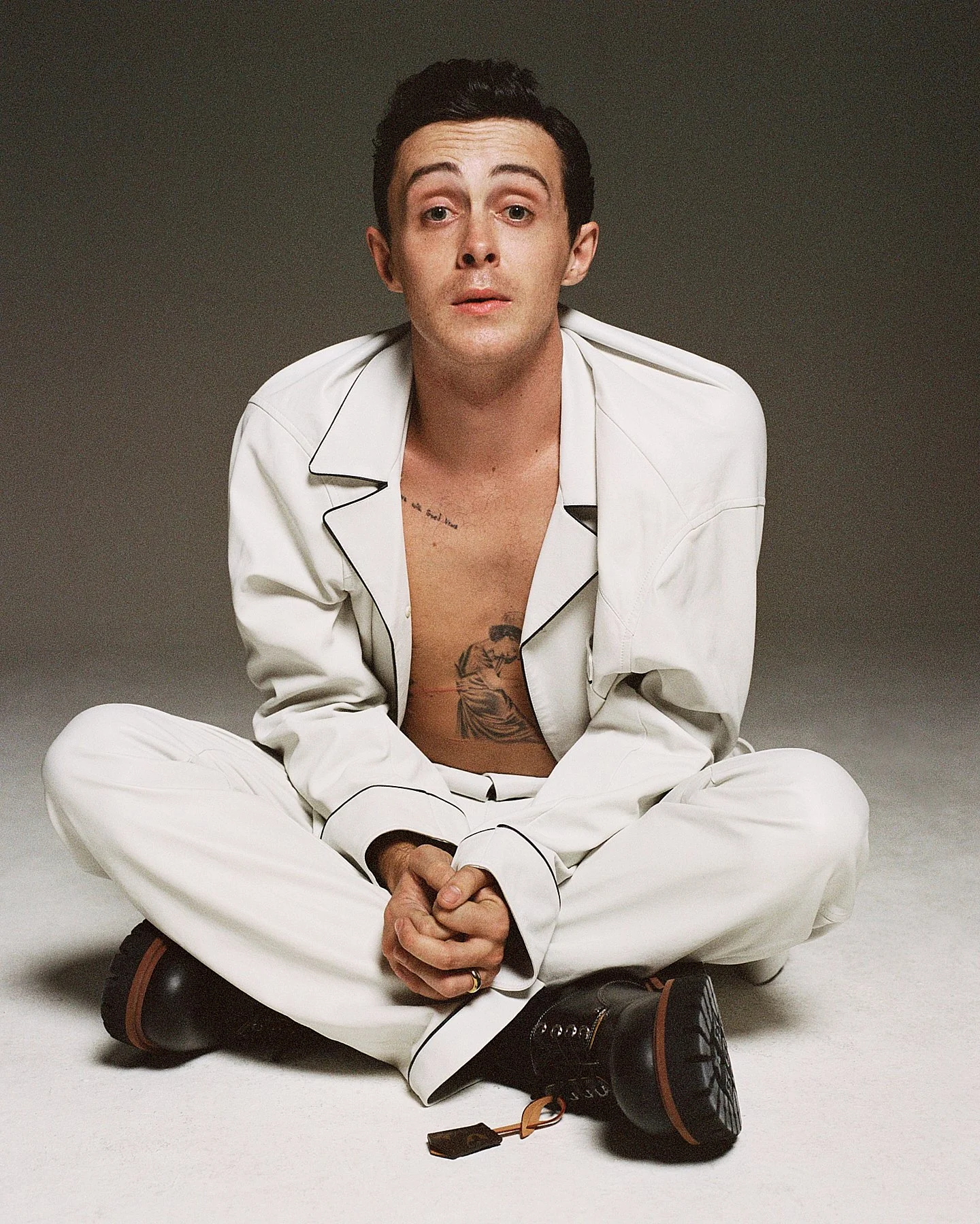 A man sitting cross-legged on the floor, wearing a white pajama set with black piping, black shoes, and a ring on his finger. He has dark hair and tattoos on his chest and shoulder and is looking directly at the camera with a neutral expression.