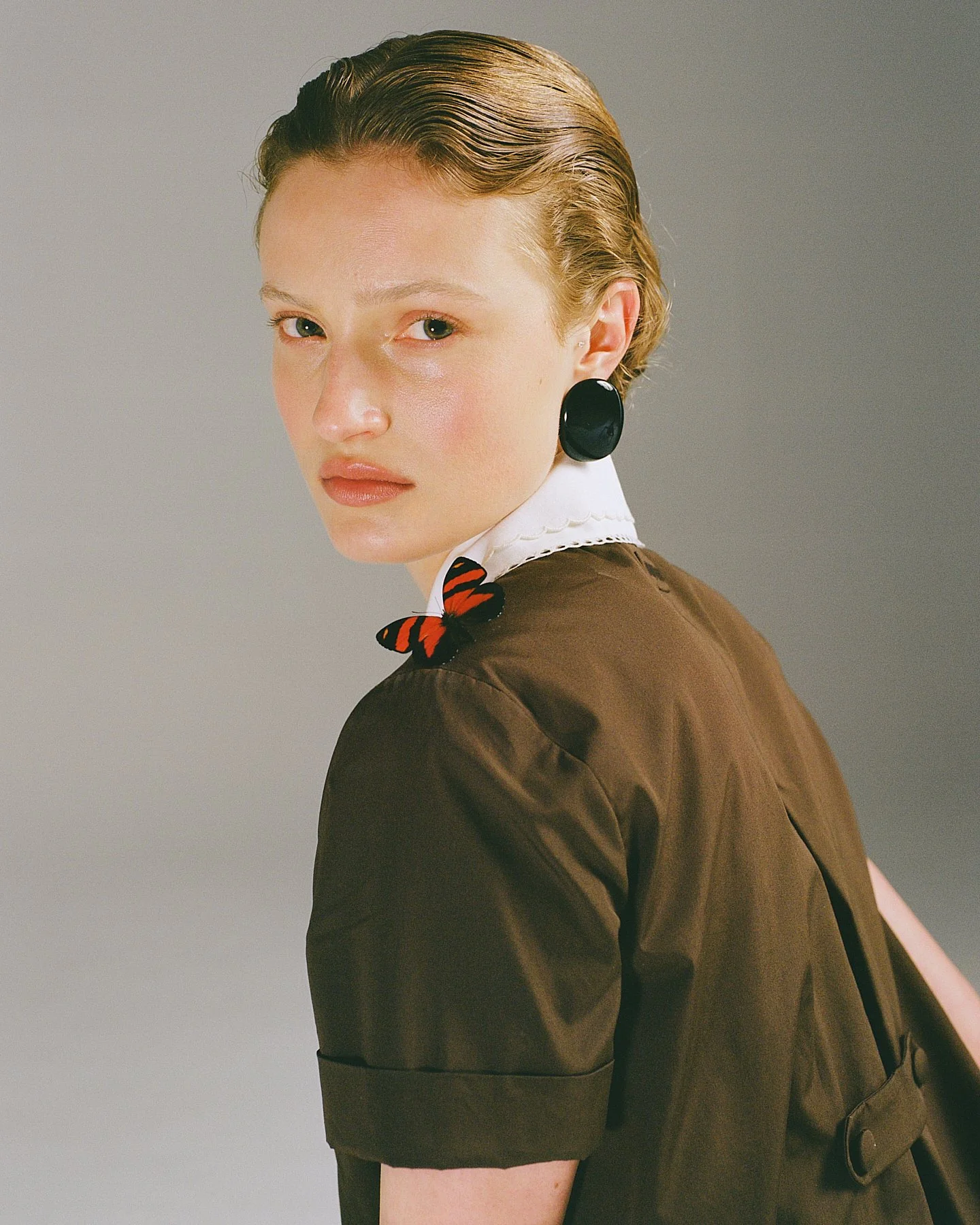 Close-up of a woman with short, wet-looking hair, wearing large black earrings, a brown jacket, and a white collared shirt with a red and black butterfly brooch.