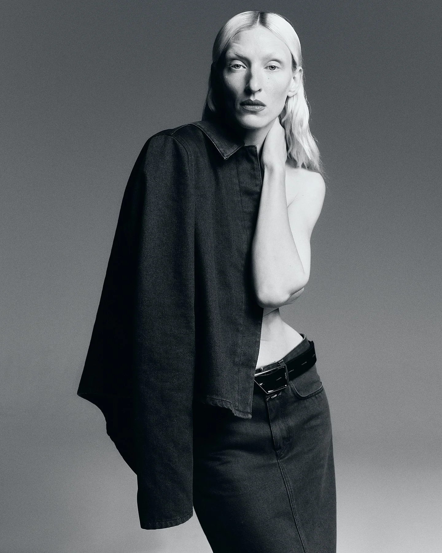 A black and white photo of a woman with platinum blonde hair, wearing a dark blazer and dark jeans, posing with her hand on her neck against a plain background.