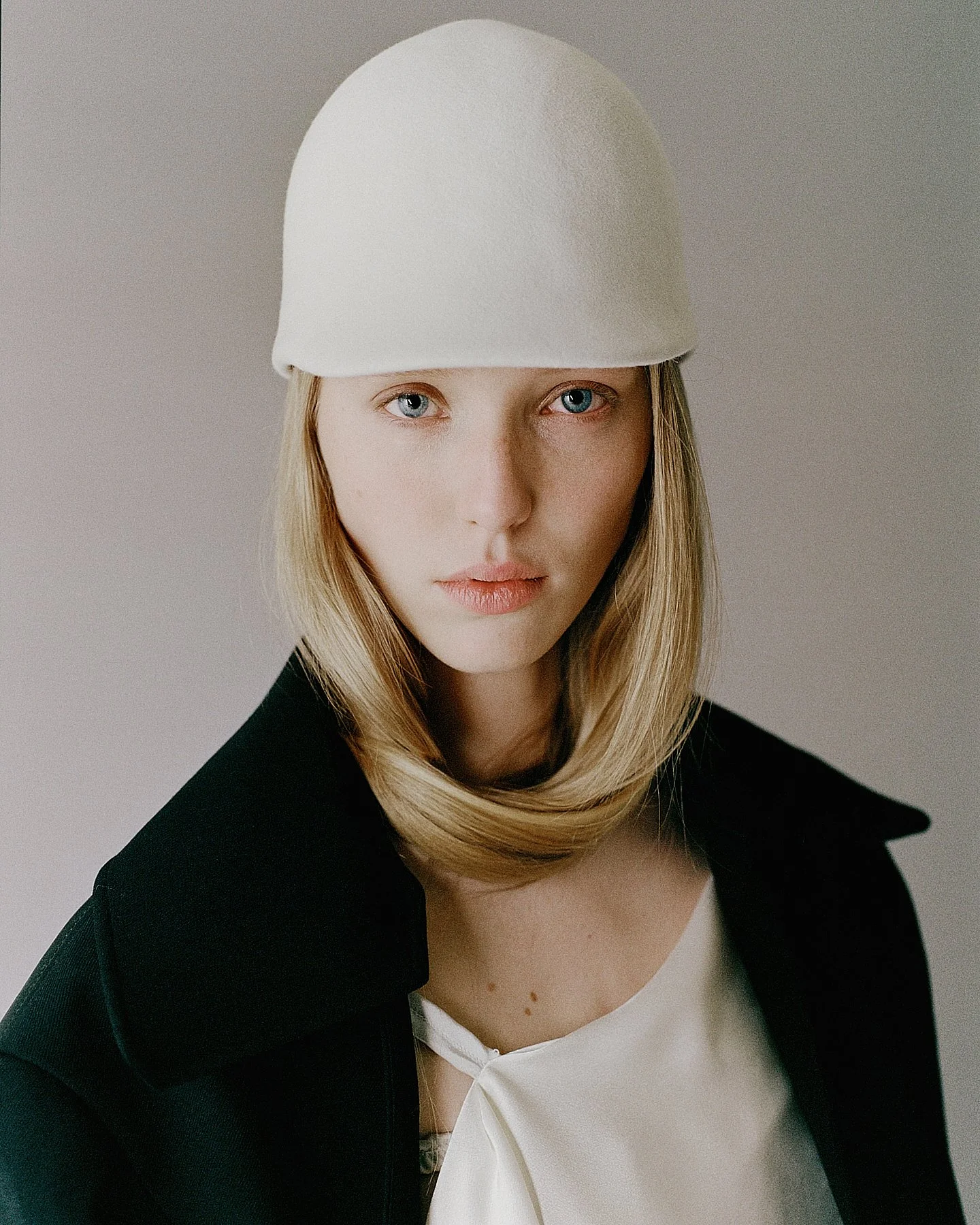 A young woman with blonde hair wearing a white hat, black blazer, and white top, looking directly at the camera against a neutral background.