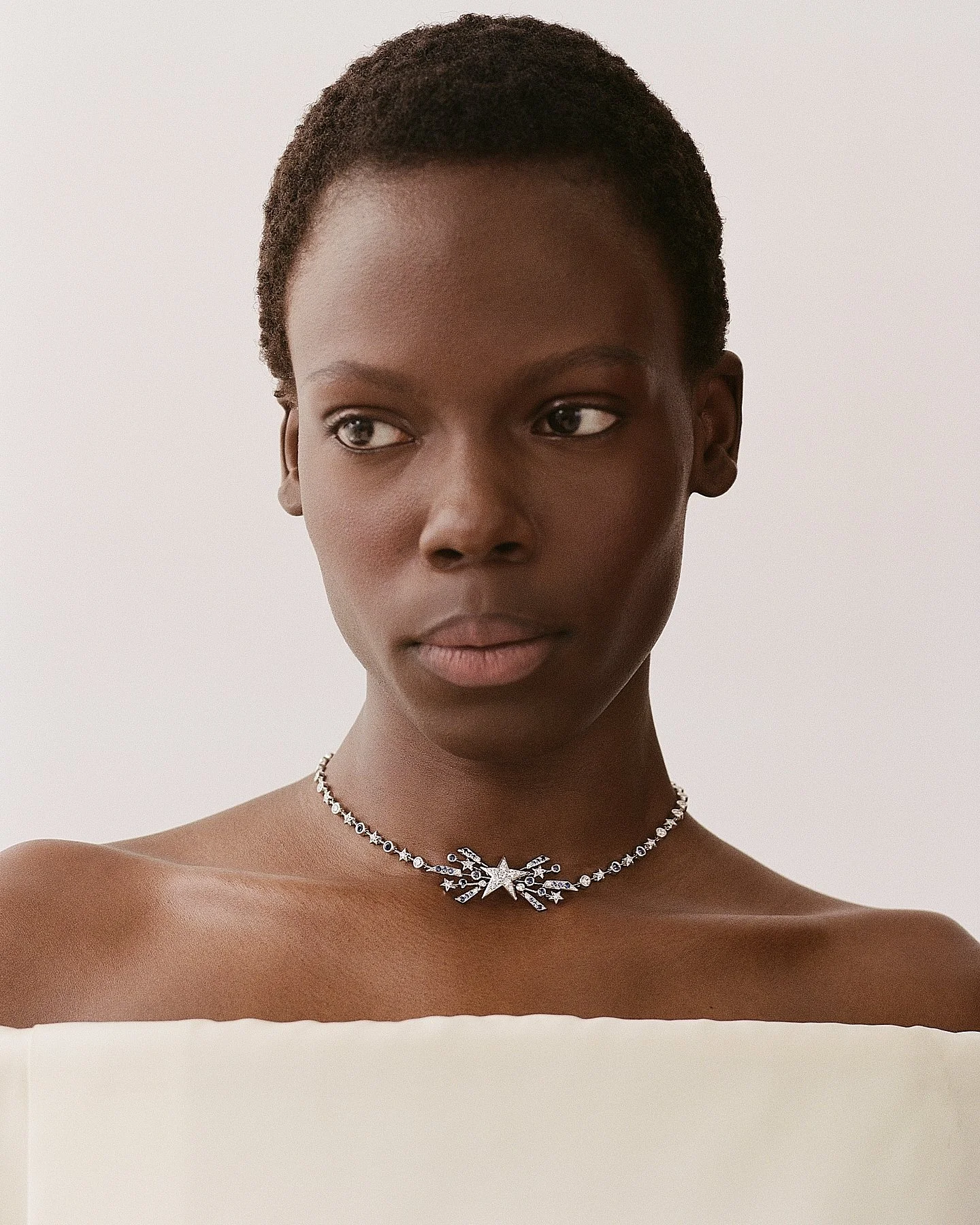 A woman with short, natural hair and dark skin wearing a silver star-shaped necklace with smaller star and moon charms. Her shoulders are bare against a plain white background.