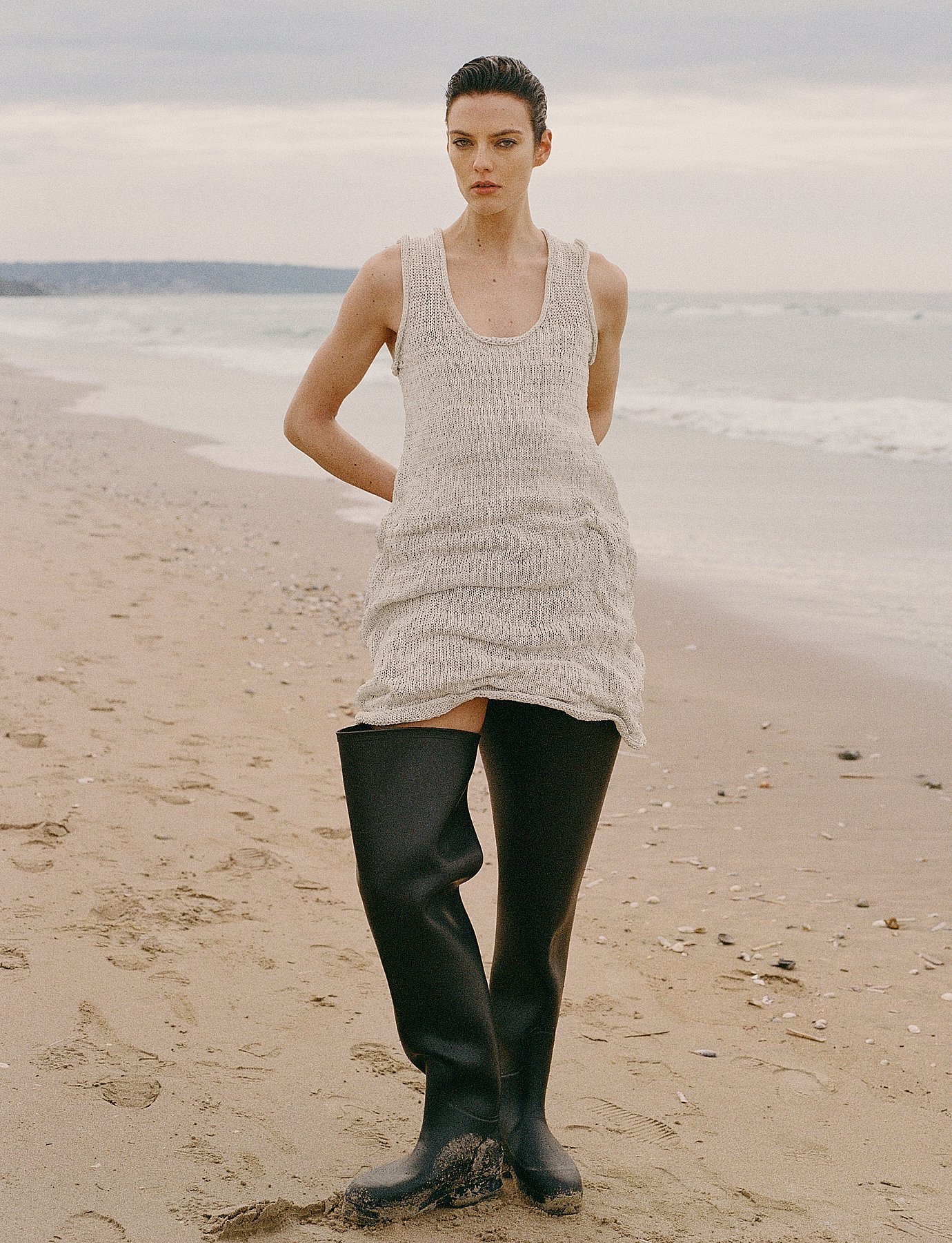 A woman standing on a sandy beach wearing a beige knitted dress and black thigh-high boots.