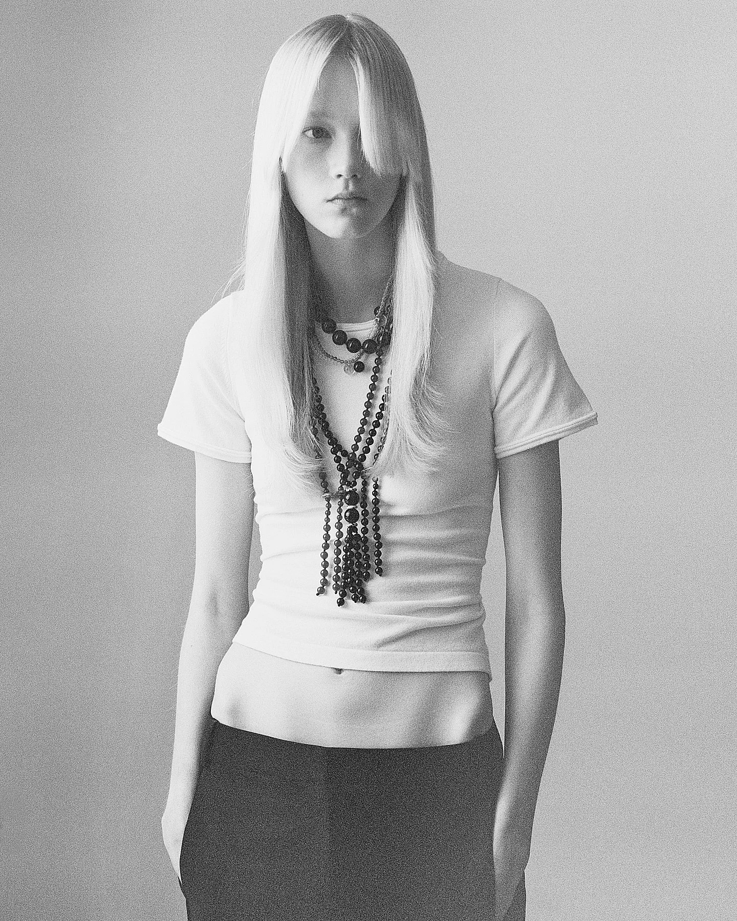 Black and white photo of a young woman with long, straight hair, wearing a white t-shirt and multiple black beaded necklaces, standing against a plain wall.
