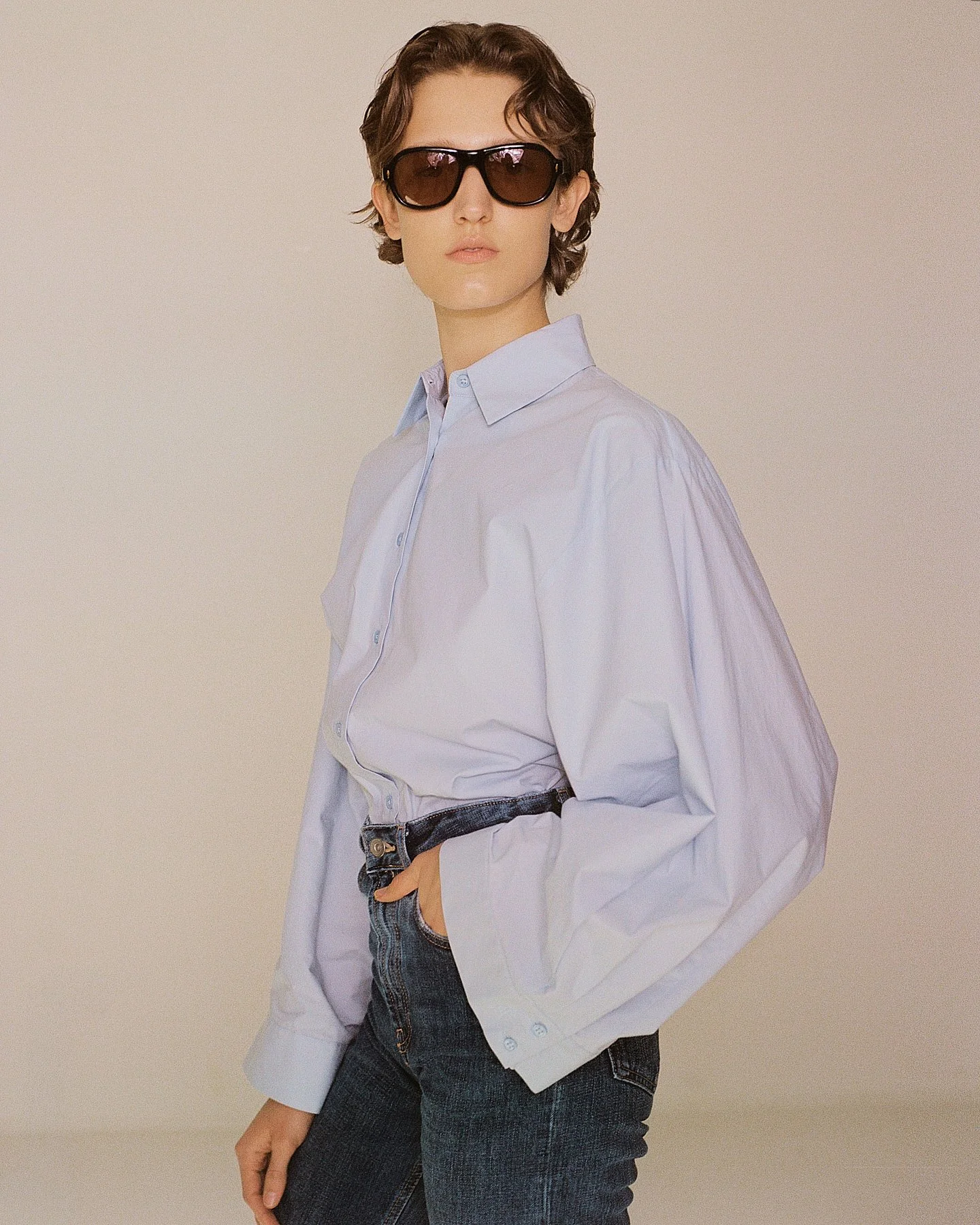 A person wearing a white oversized shirt, dark jeans, and sunglasses posing against a plain background.