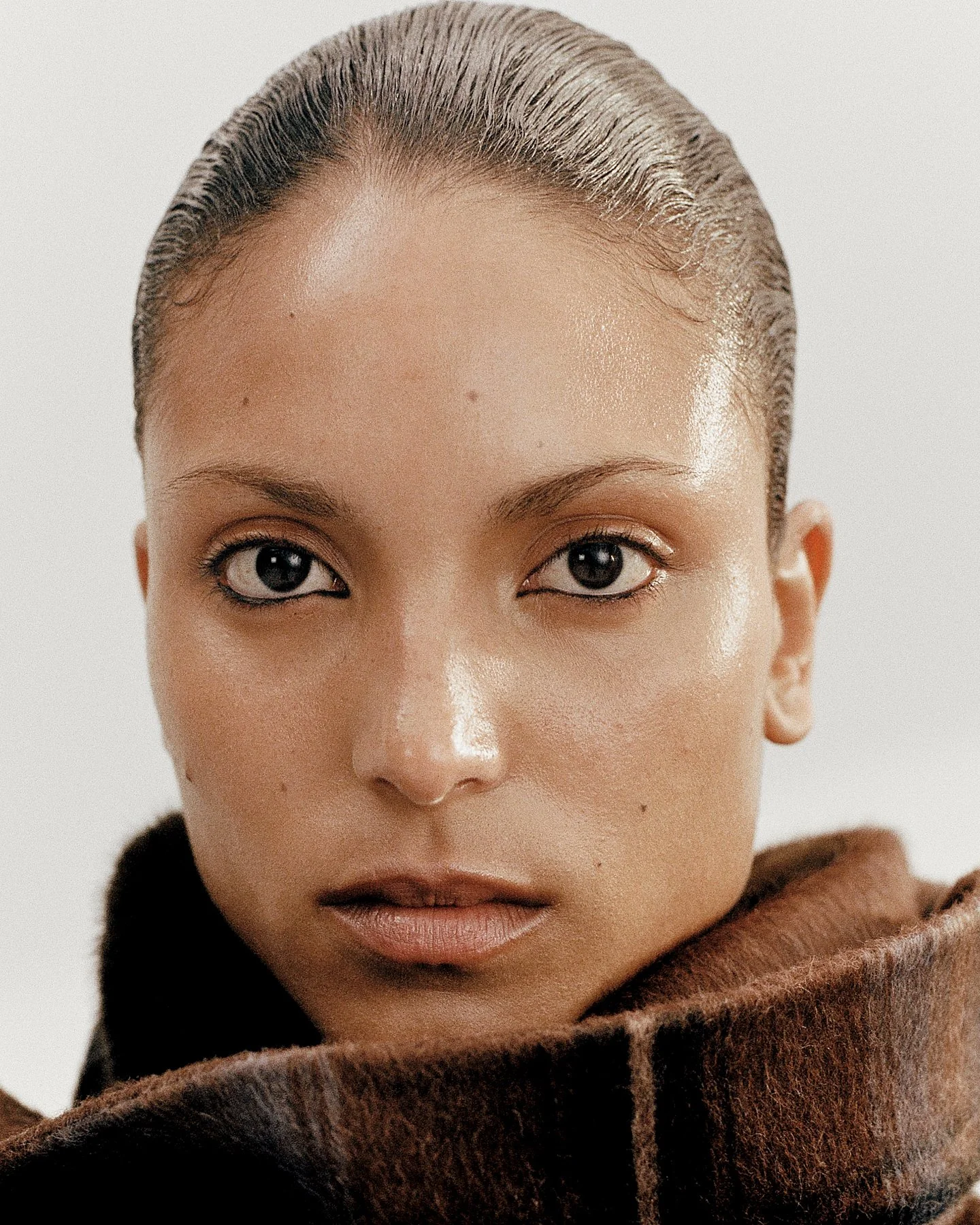 Close-up of a person's face with slicked-back hair, dark eyes, and brown skin, wearing a brown fur coat