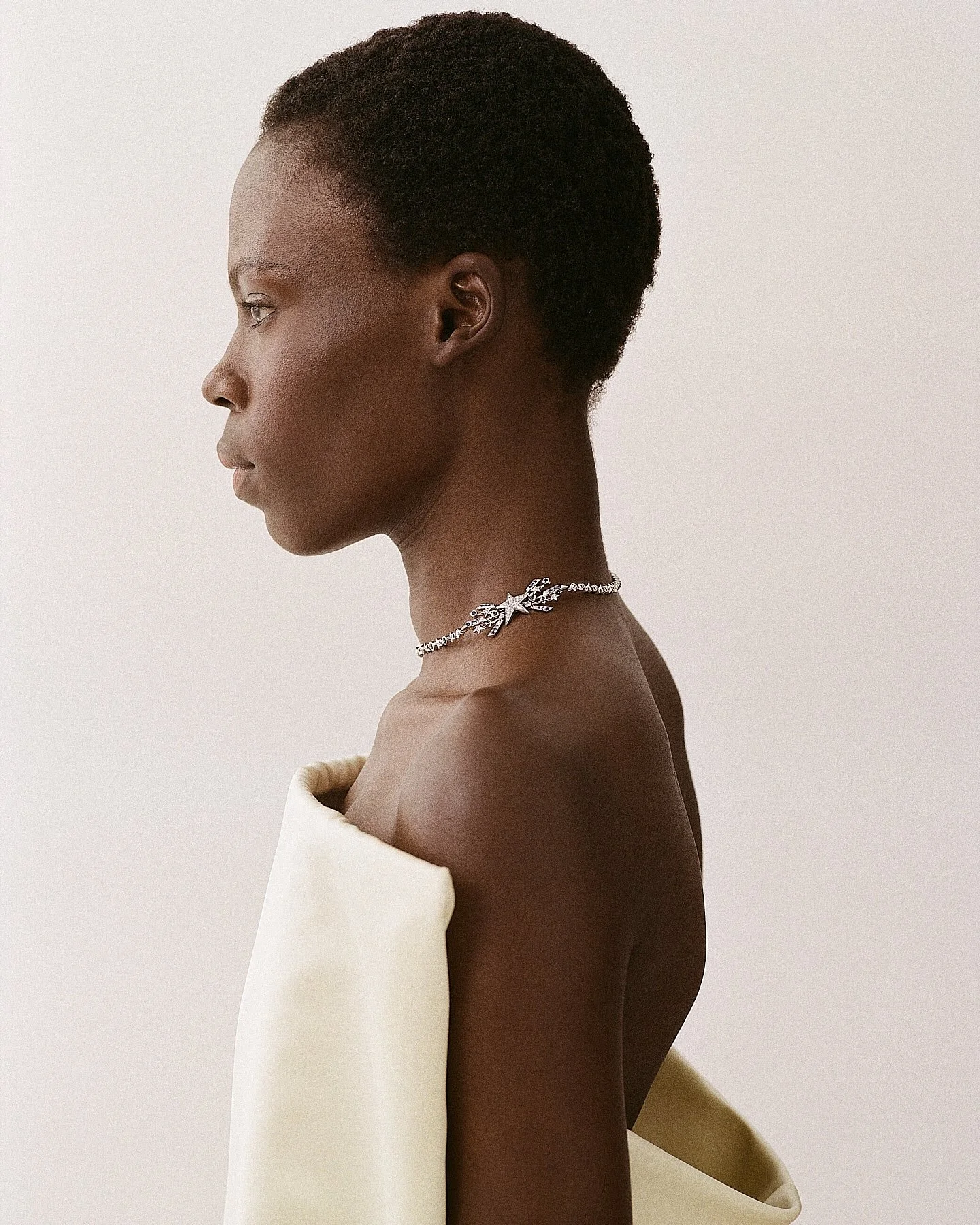 Profile of a woman with short natural hair wearing a silver star-shaped necklace and a cream-colored off-the-shoulder top against a plain white background.