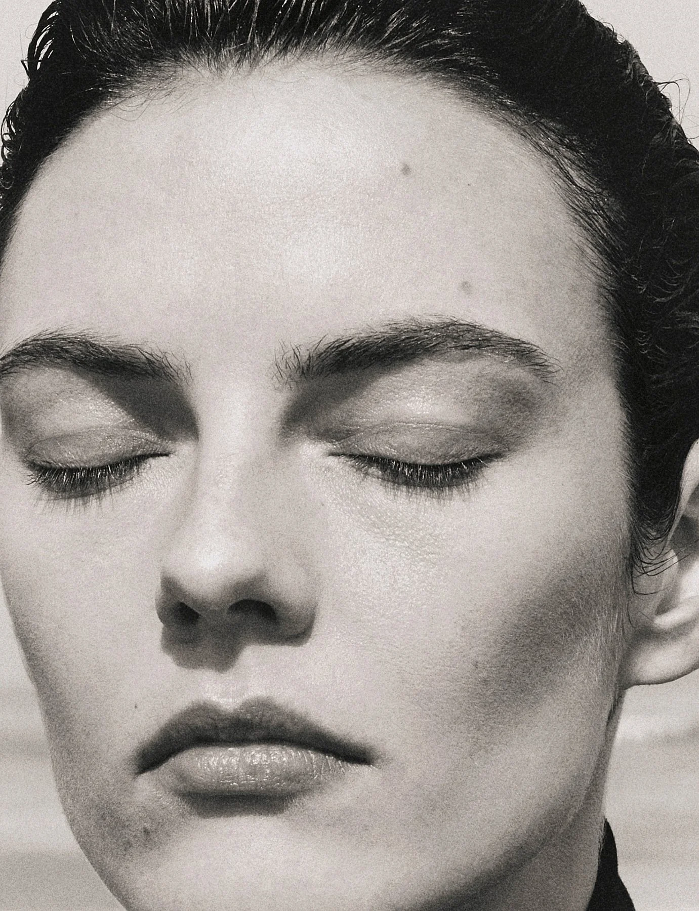 Close-up black and white photograph of a woman with her eyes closed, showing her eyebrows, eyelashes, nose, and lips.