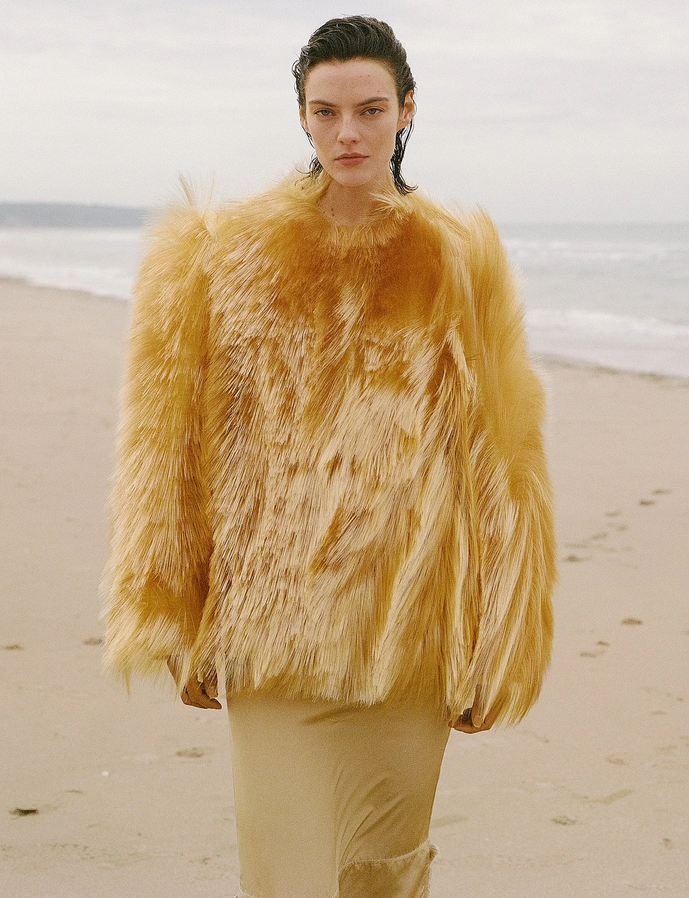 A woman with dark wet hair and light skin wearing a thick, yellow faux fur coat and a light-colored dress, standing on a sandy beach with the ocean and sky in the background.