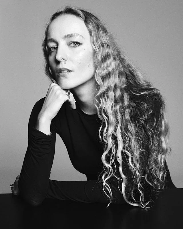 Black and white portrait of a woman with long, wavy hair resting her chin on her hand, wearing a dark top, looking at the camera.