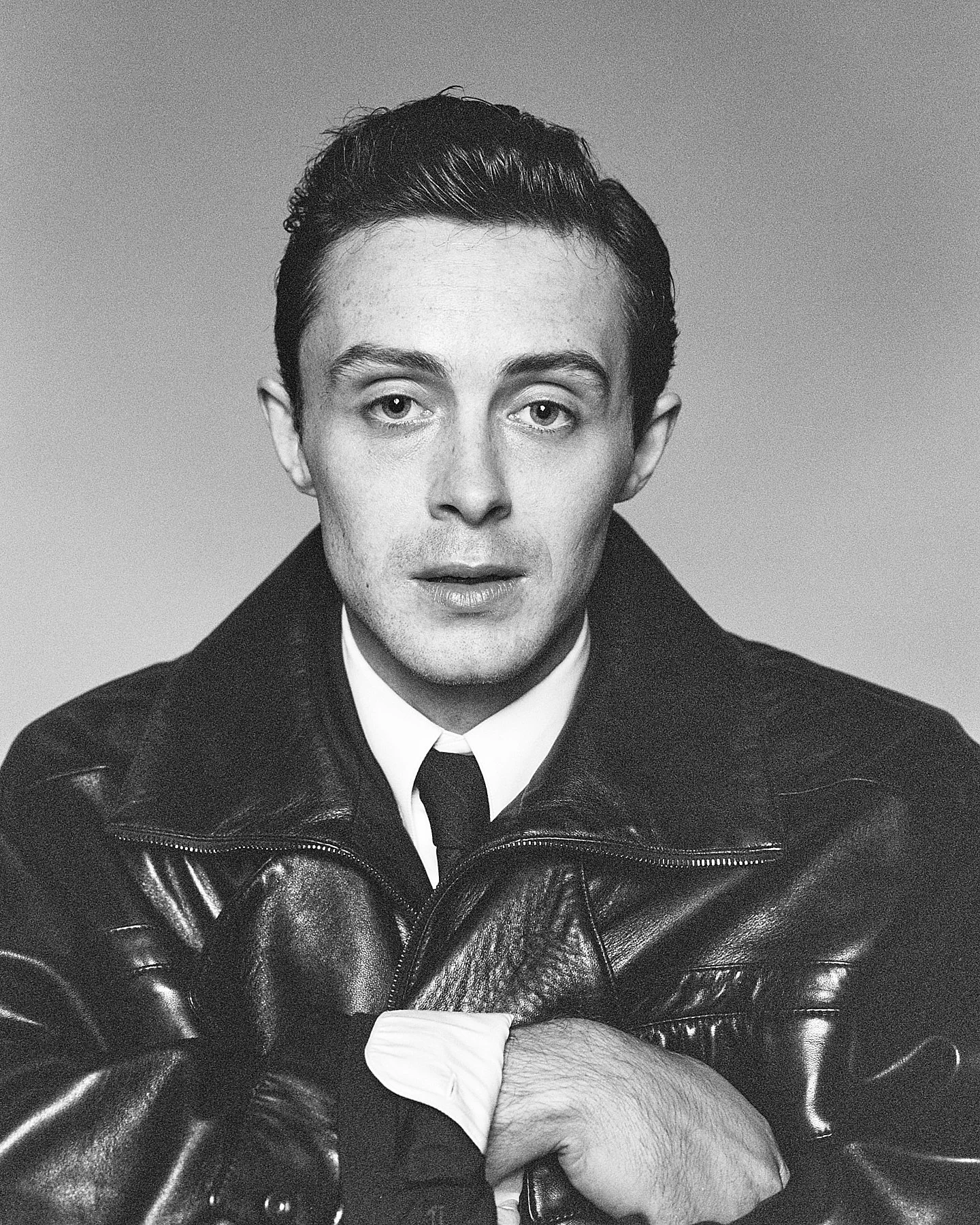 Black and white portrait of a young man with dark, slicked-back hair, wearing a white shirt, black tie, and a leather jacket. He is looking directly at the camera with a neutral expression, and his hands are crossed in front of him.