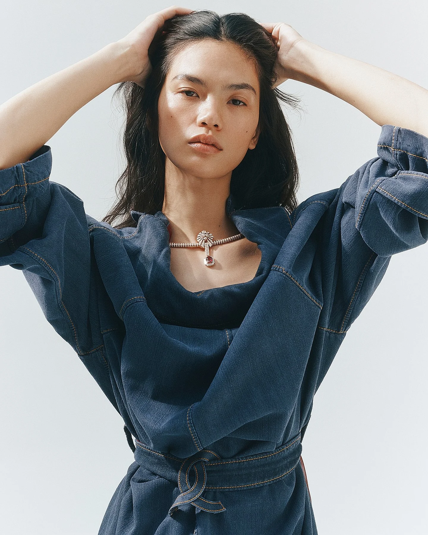 A woman with dark hair, wearing a dark denim jacket and matching jeans, posing with her hands resting on her head against a plain light background.