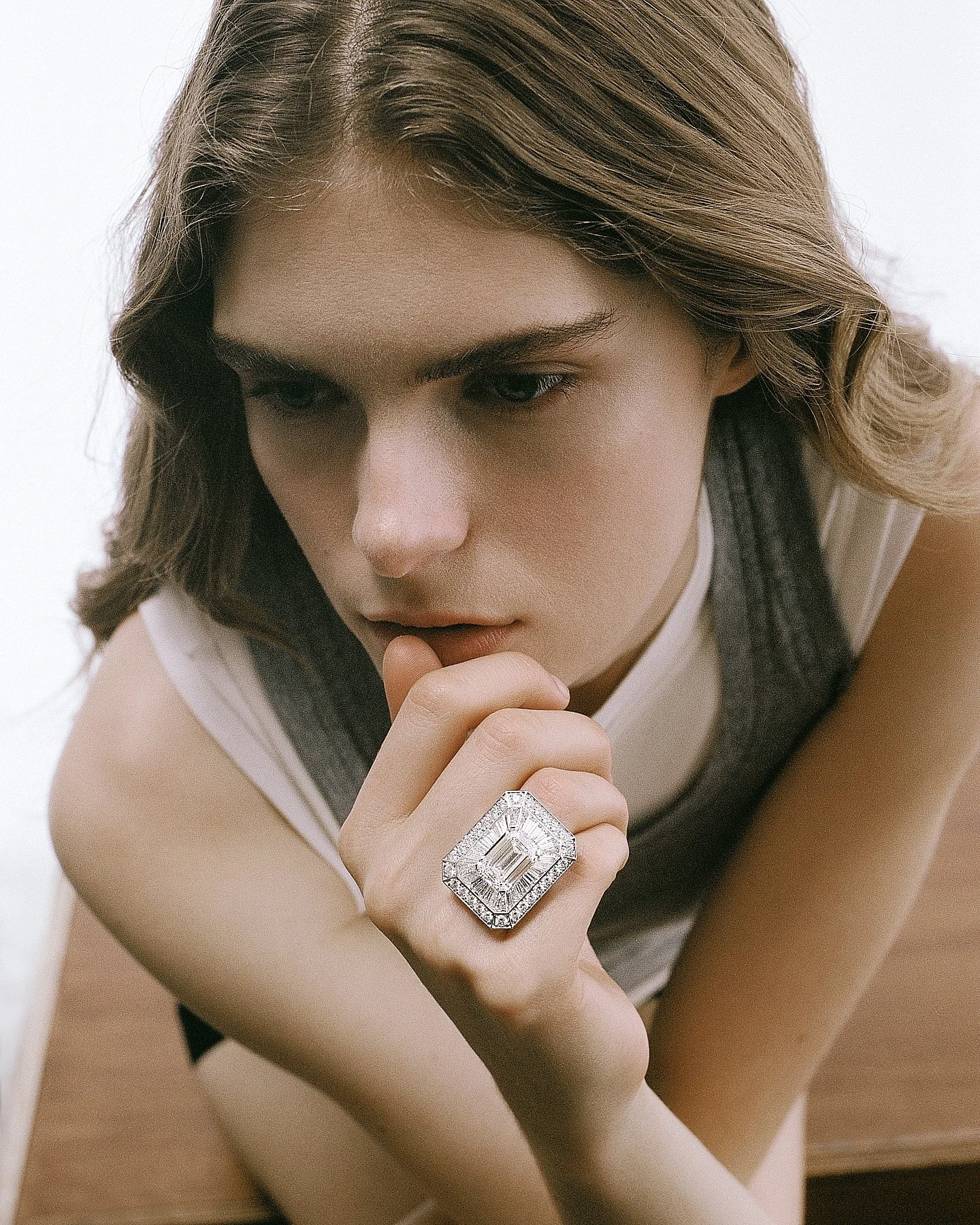 Close-up of a woman with wavy brown hair wearing a large, rectangular diamond ring on her finger. She is touching her chin with her hand and looking downward. The background is white.