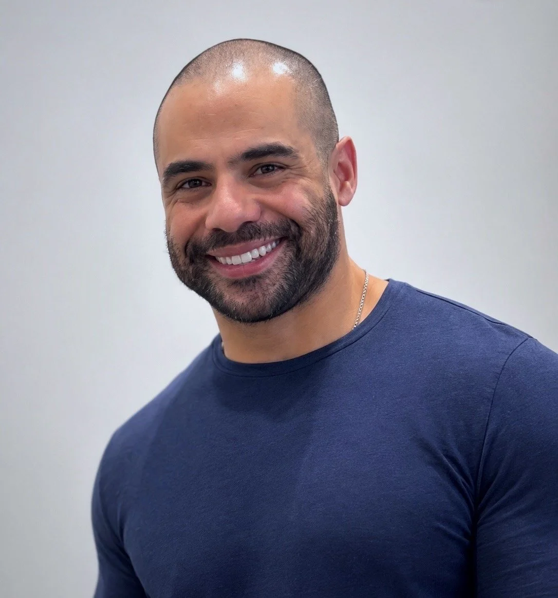 Close up of Freddy Pacheco, with a shaved head, beard, wearing a navy blue t-shirt, standing against a plain light grey background. Live Oak Renovations offers Austin sustainable, professional renovation and homebuilding.