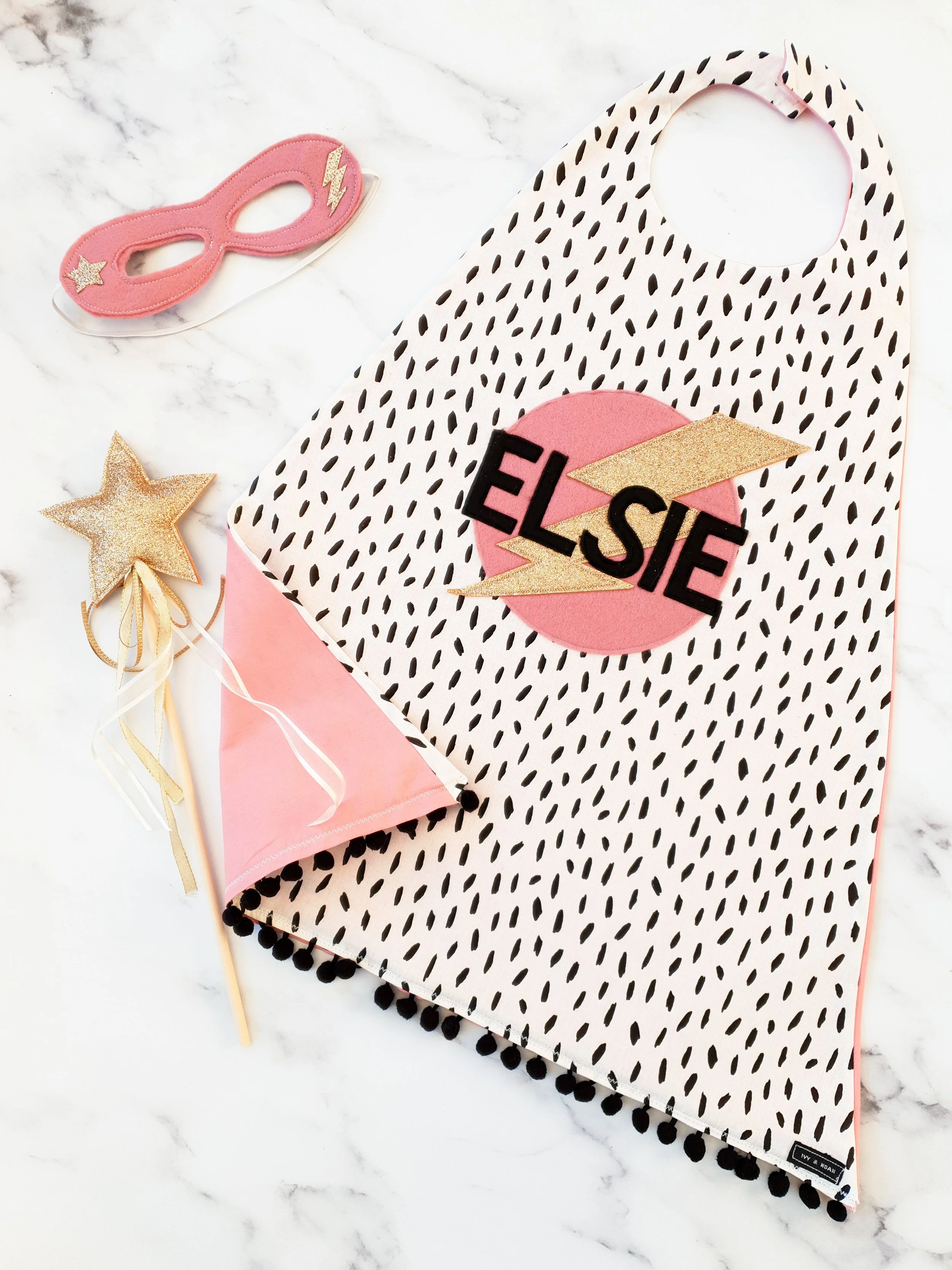 Flat lay of monochrome and pink personalised cape, mask and wand set.