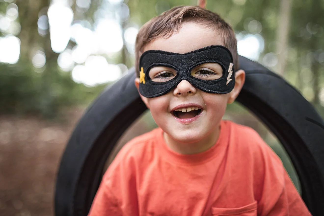 Young boy swinging on a tyre in the woods wearing a superhero mask.