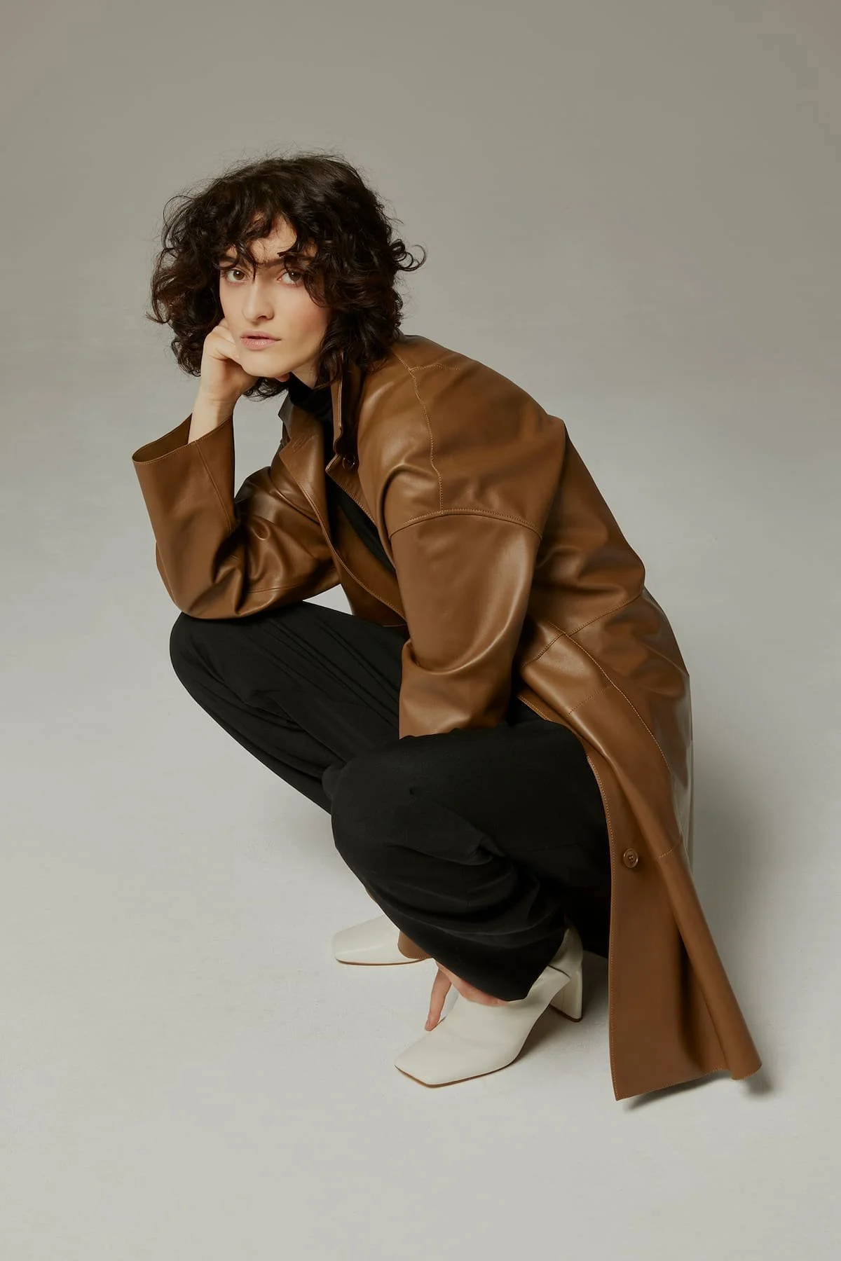 Fashion editorial styling with brown leather jacket, black trousers and sculptural pose.