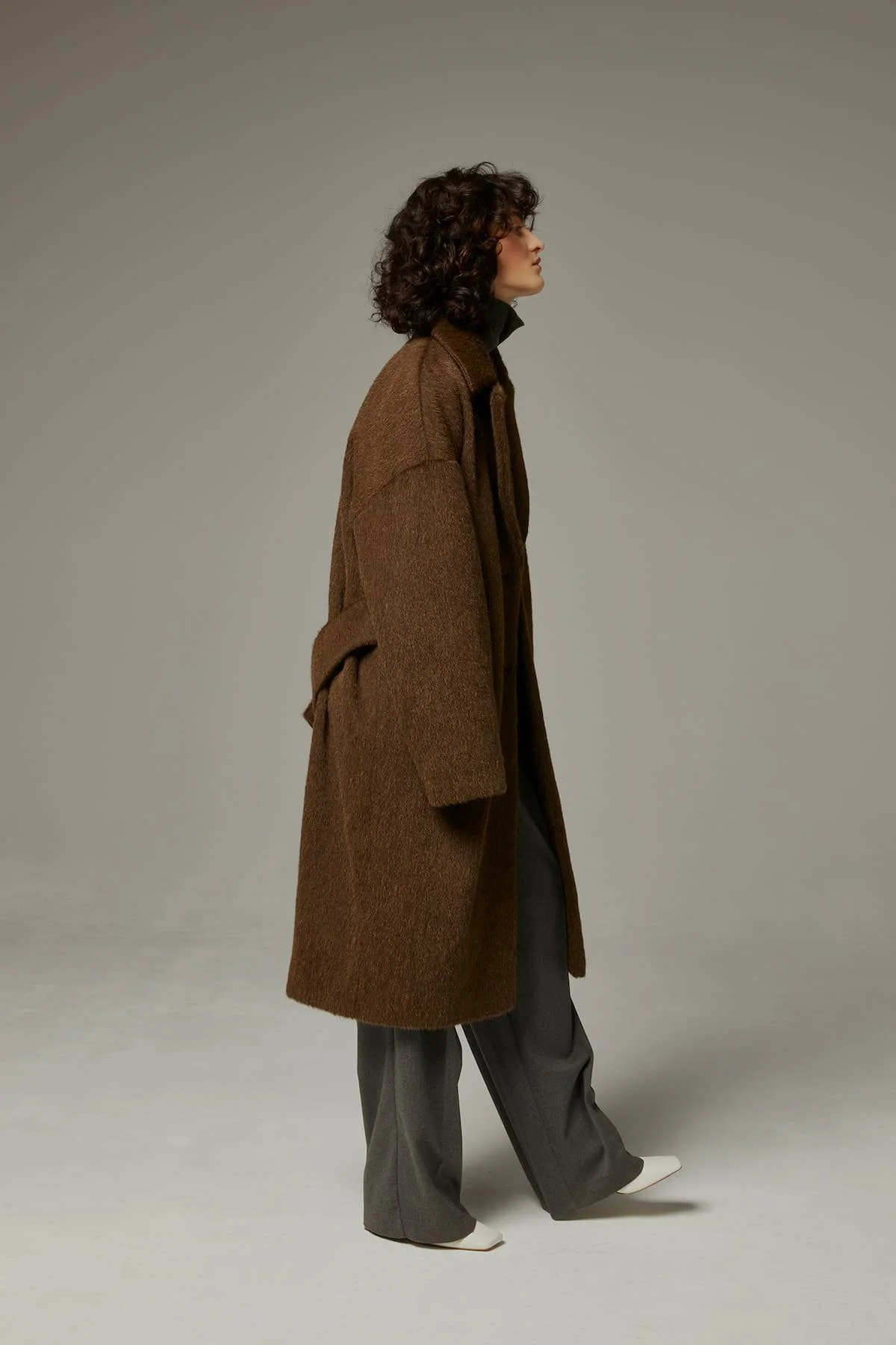 Fashion editorial styling with long brown cashmere coat, wide trousers and minimalist studio background.