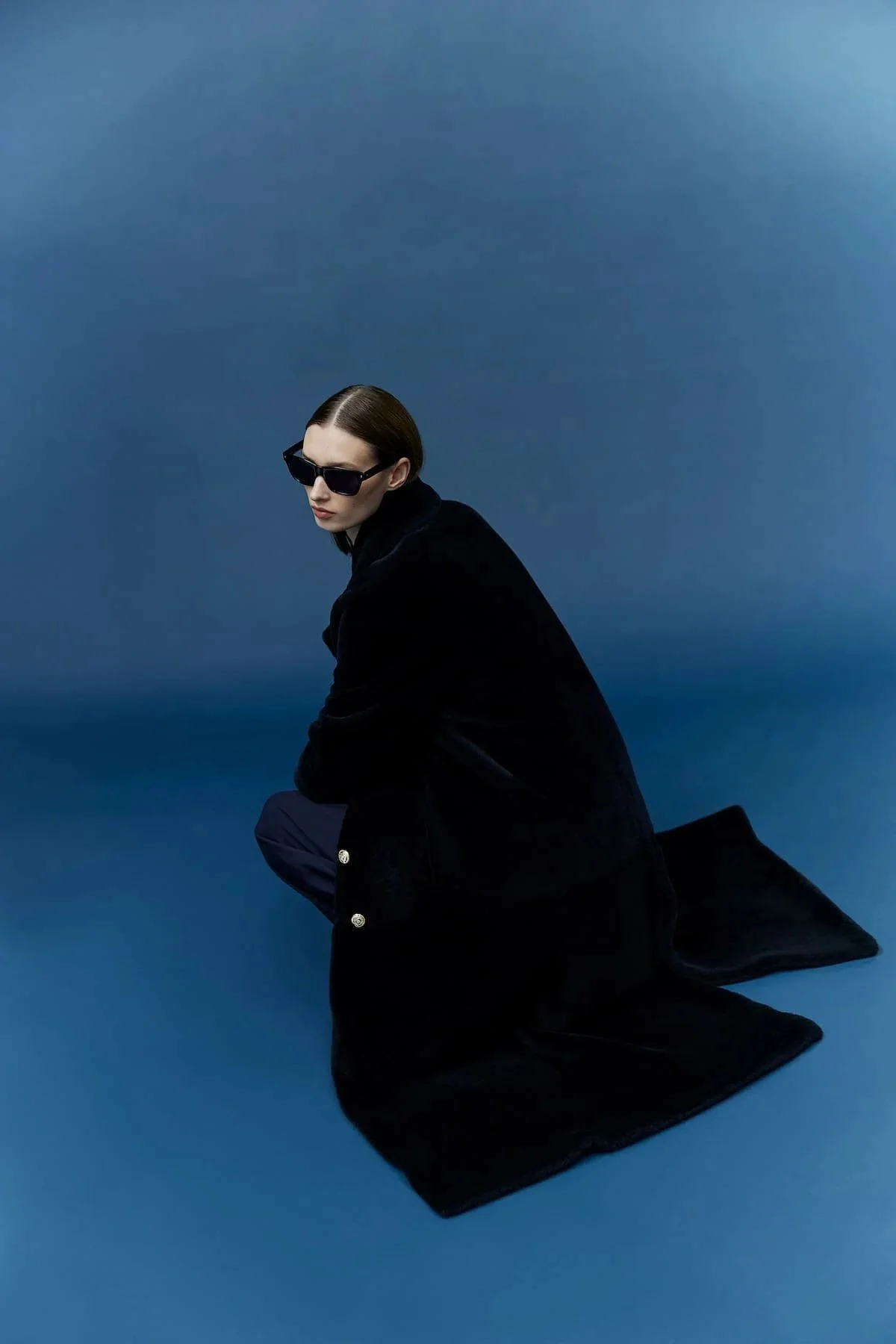 Fashion editorial image with black coat, sunglasses and deep blue studio background.