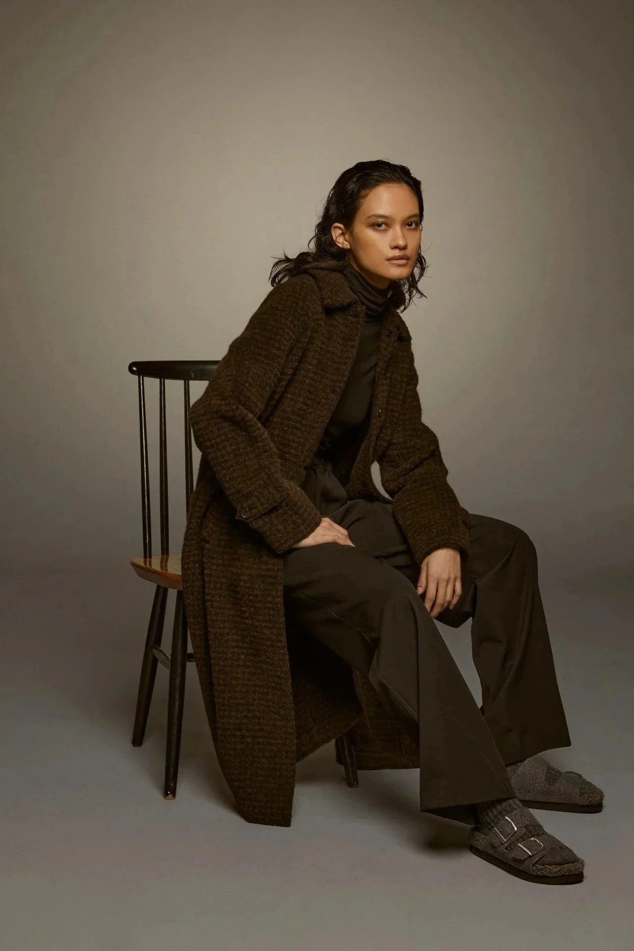 Fashion editorial image with brown textured coat, seated model and refined studio styling.