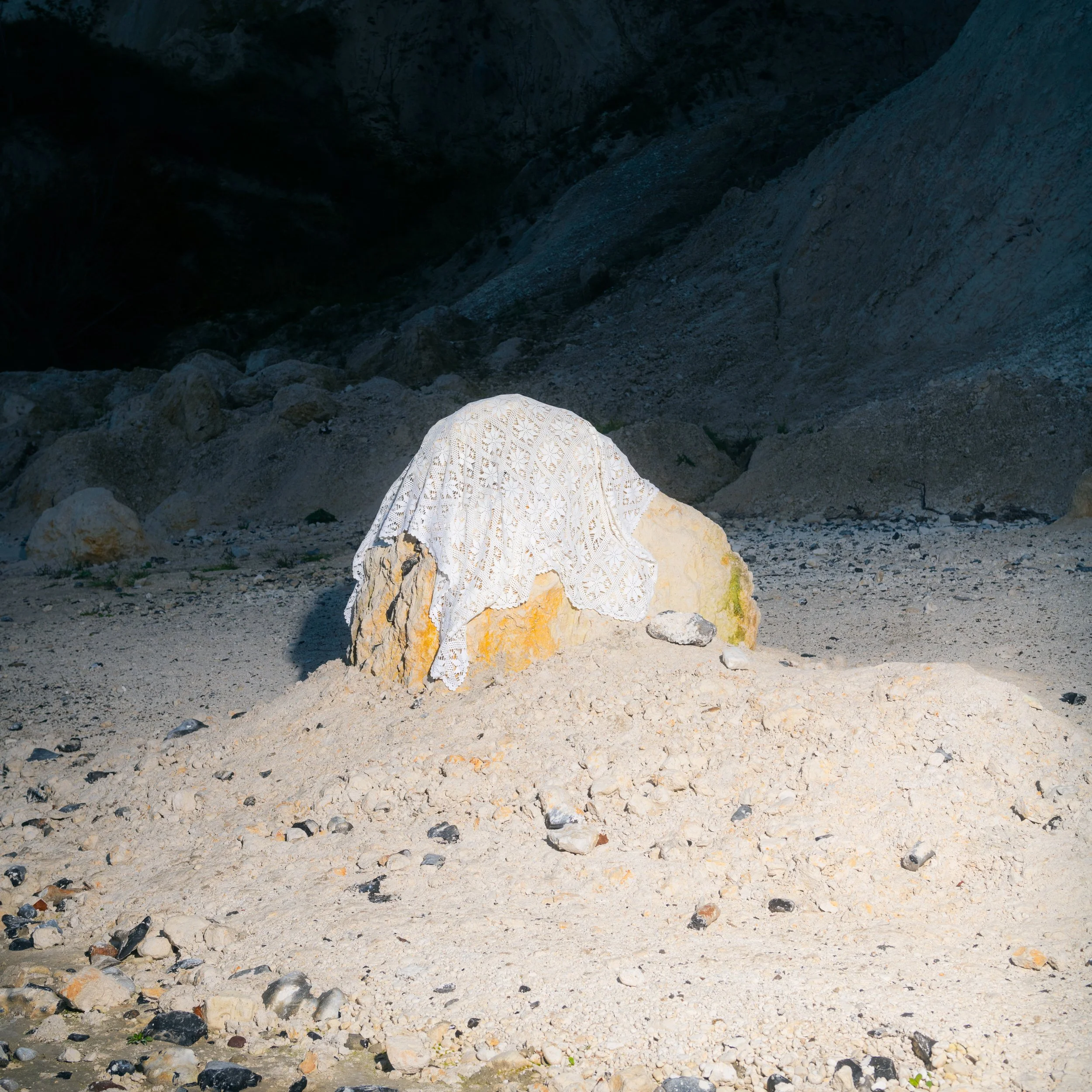 A large rock on a sandy beach, draped with a white lace cloth, with a backdrop of dark cliffs and scattered smaller rocks.
