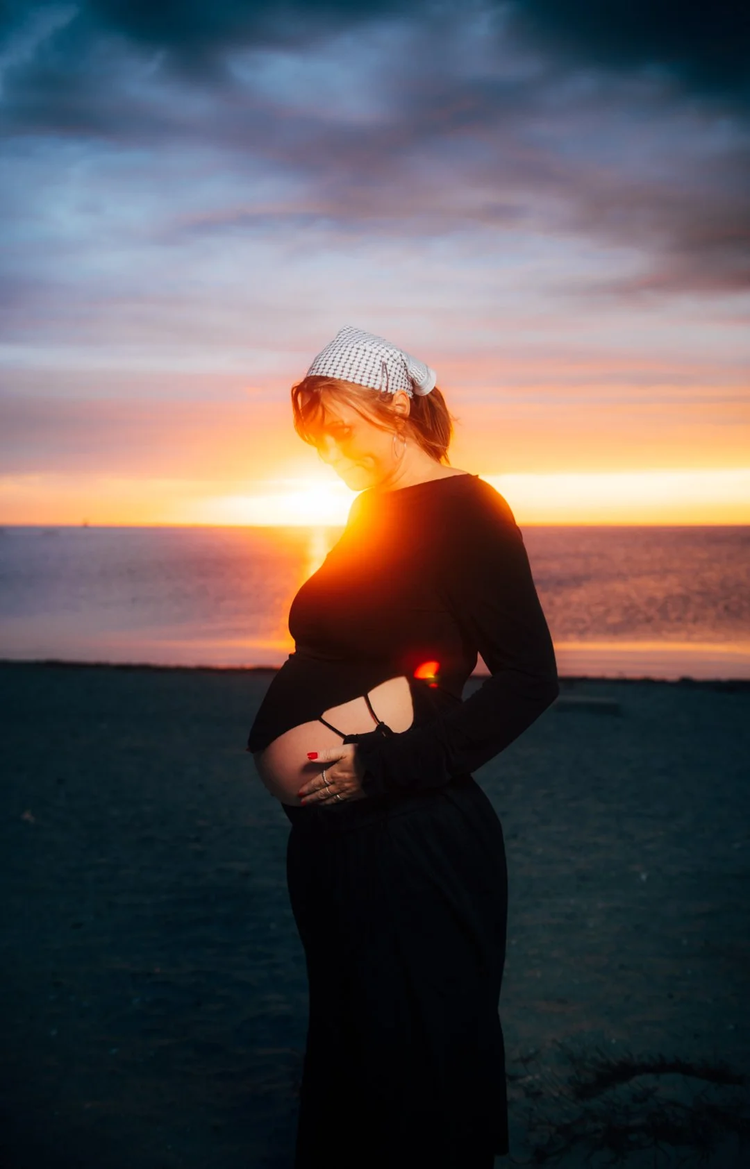 Pregnant woman standing on the beach at sunset, holding her belly, with the sun setting over the ocean in the background.