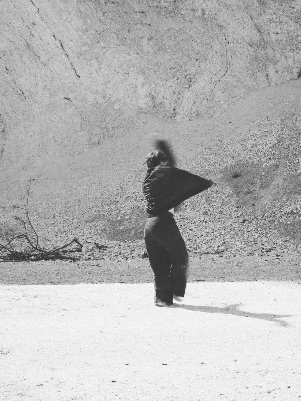 Person standing in sandy outdoor area near rocky wall, captured in black and white, with blurred motion.