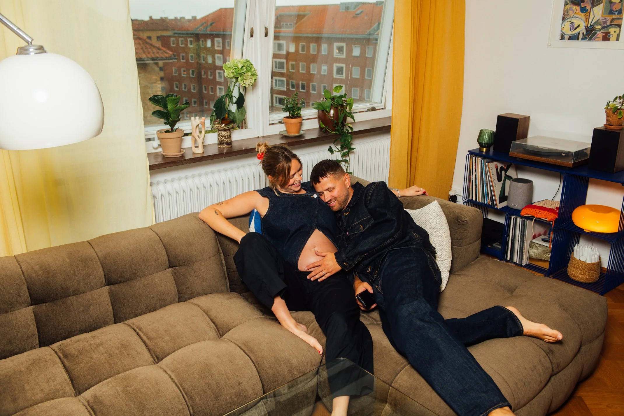 A pregnant woman and a man are sitting on a beige sofa indoors. The woman is smiling and has her shirt lifted to reveal her pregnant belly. The man is leaning over and touching her belly. There are houseplants on the windowsill behind them and a window showing apartment buildings outside.