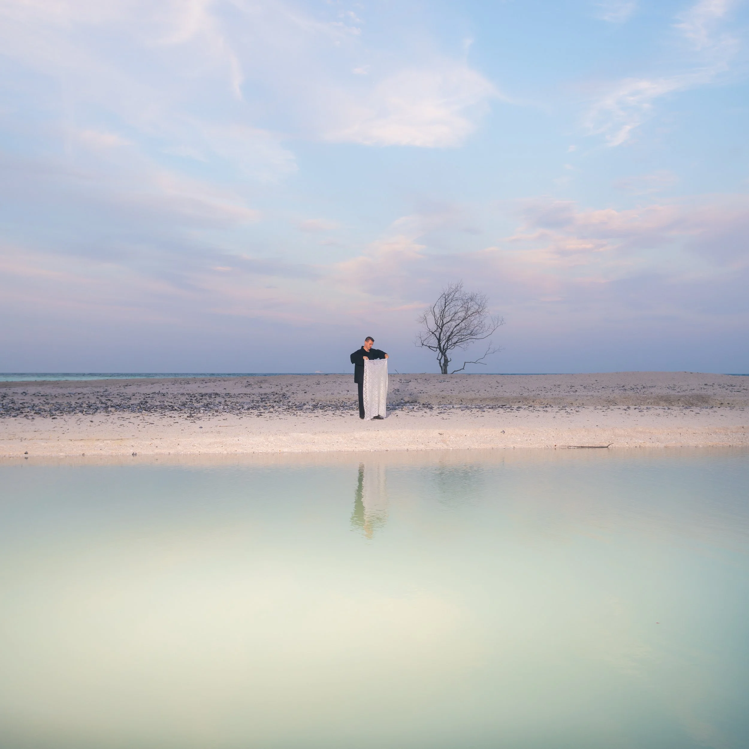 A person in a black jacket standing on a sandy beach near a body of water, holding a long white item, with a leafless tree in the background and a cloudy sky overhead.