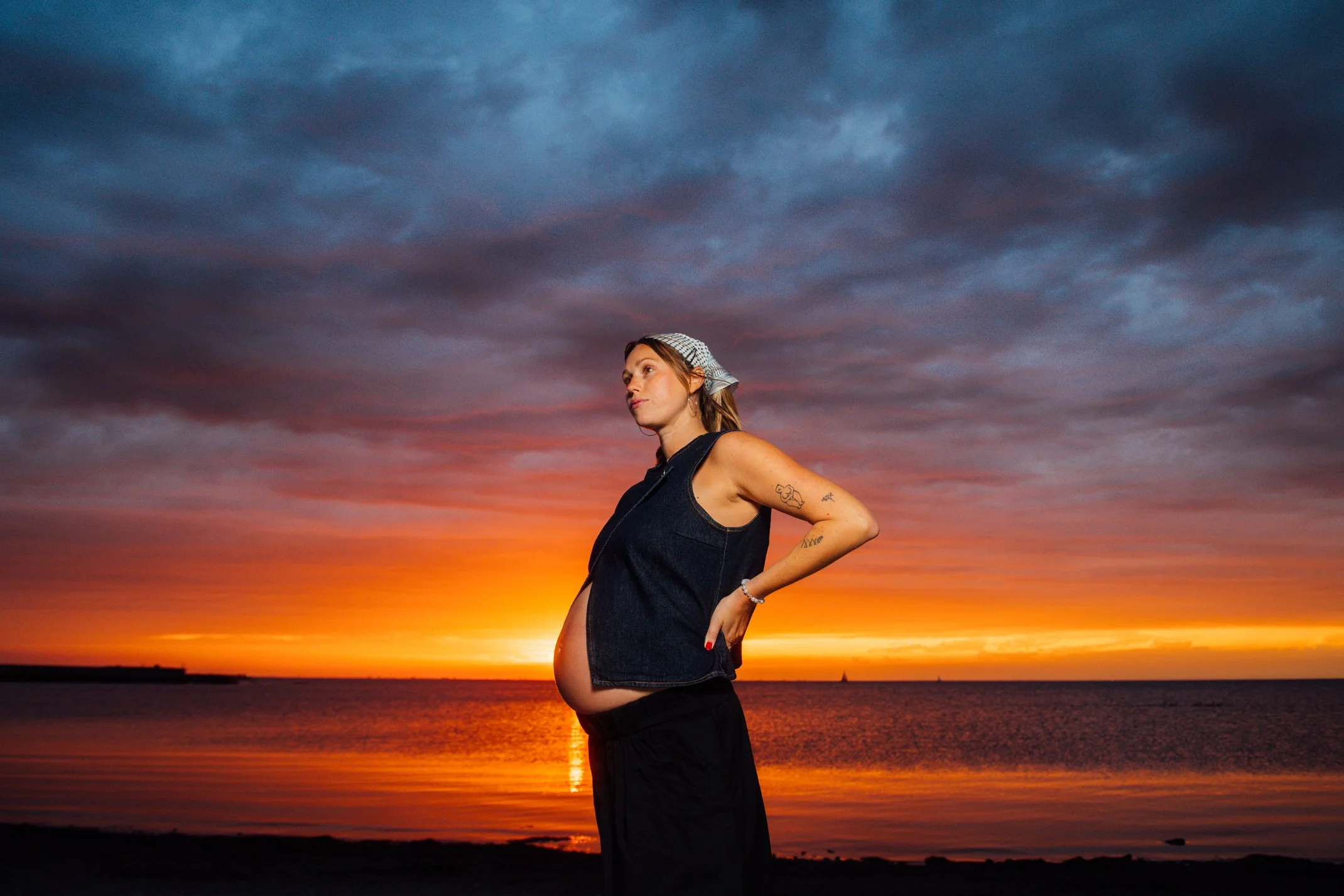 A pregnant woman stands by a body of water during sunset, wearing a sleeveless denim vest, a headscarf, and black pants, with tattoos on her arm and a bracelet on her wrist.