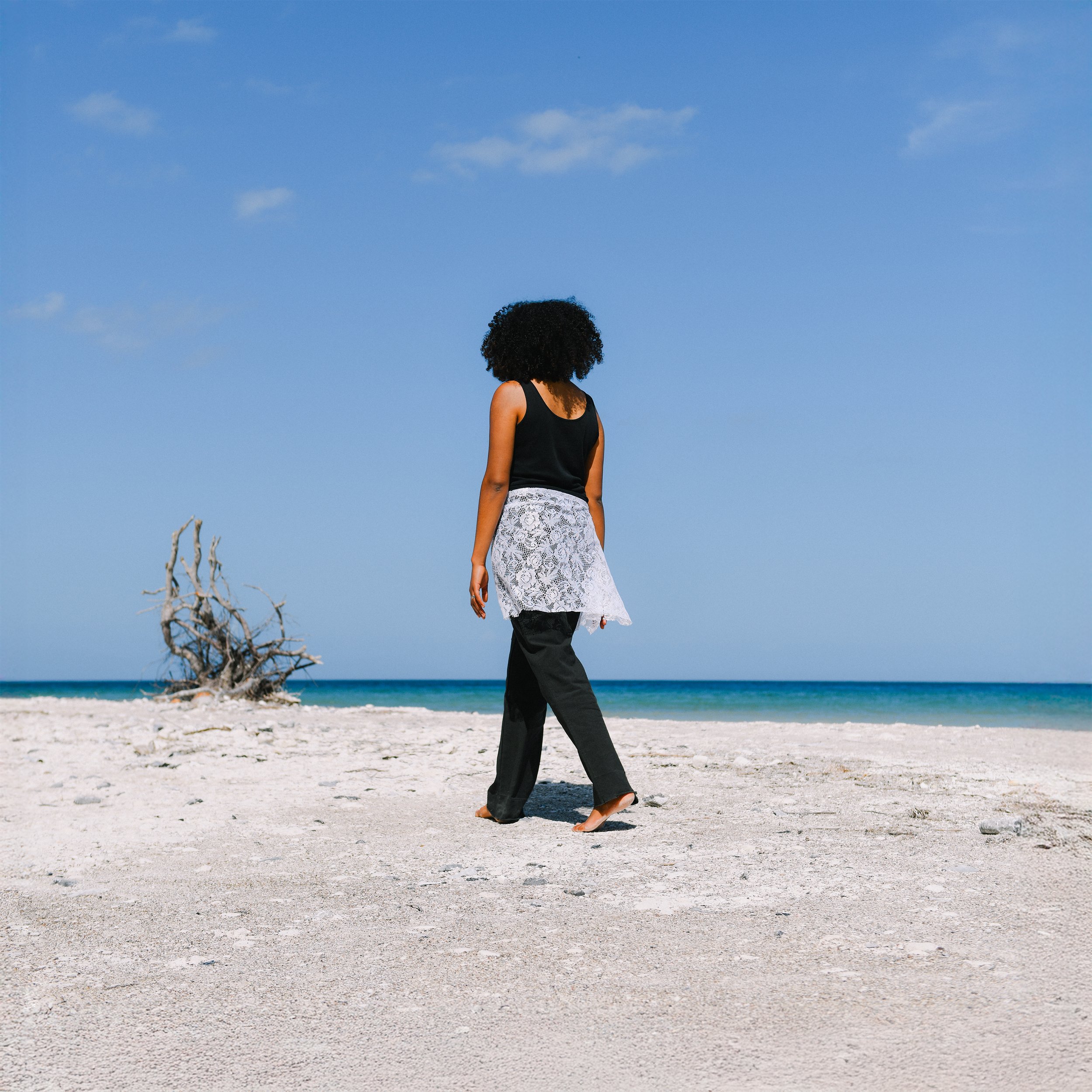 Woman in black tank top and black pants walking on sandy beach with dead tree and ocean in background