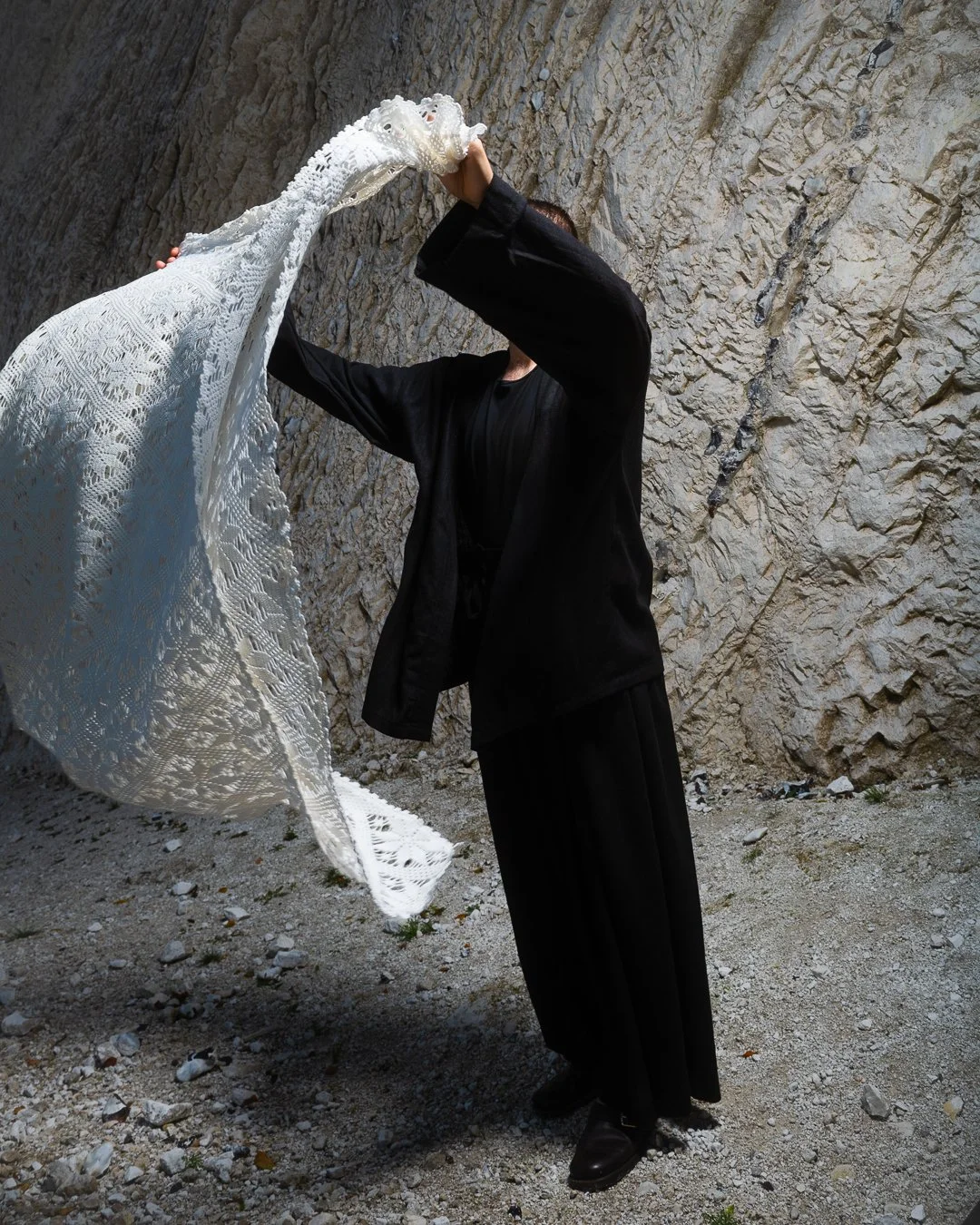 A person holding a large, white, lacy textile item against a rocky wall background.