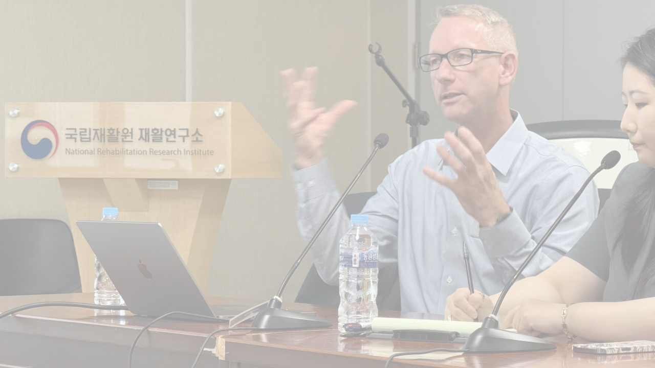 A man with glasses and a light blue shirt speaking at a panel discussion, with a woman beside him taking notes, in a room at the National Rehabilitation Research Institute, which is indicated by the sign in Korean and English on the podium.