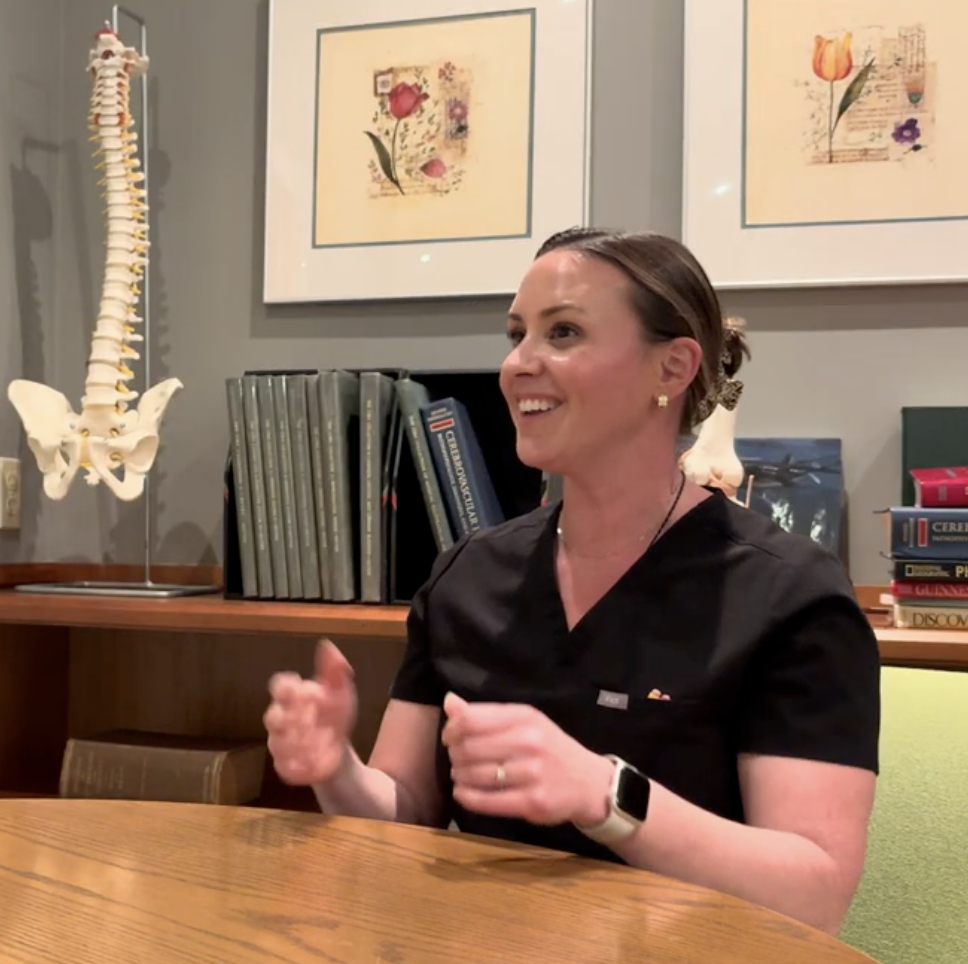 A woman with dark hair tied back, wearing a black medical uniform and a smartwatch, sitting at a wooden table and smiling. In the background, there is a model of a human spine, framed floral artwork, and shelves with books.