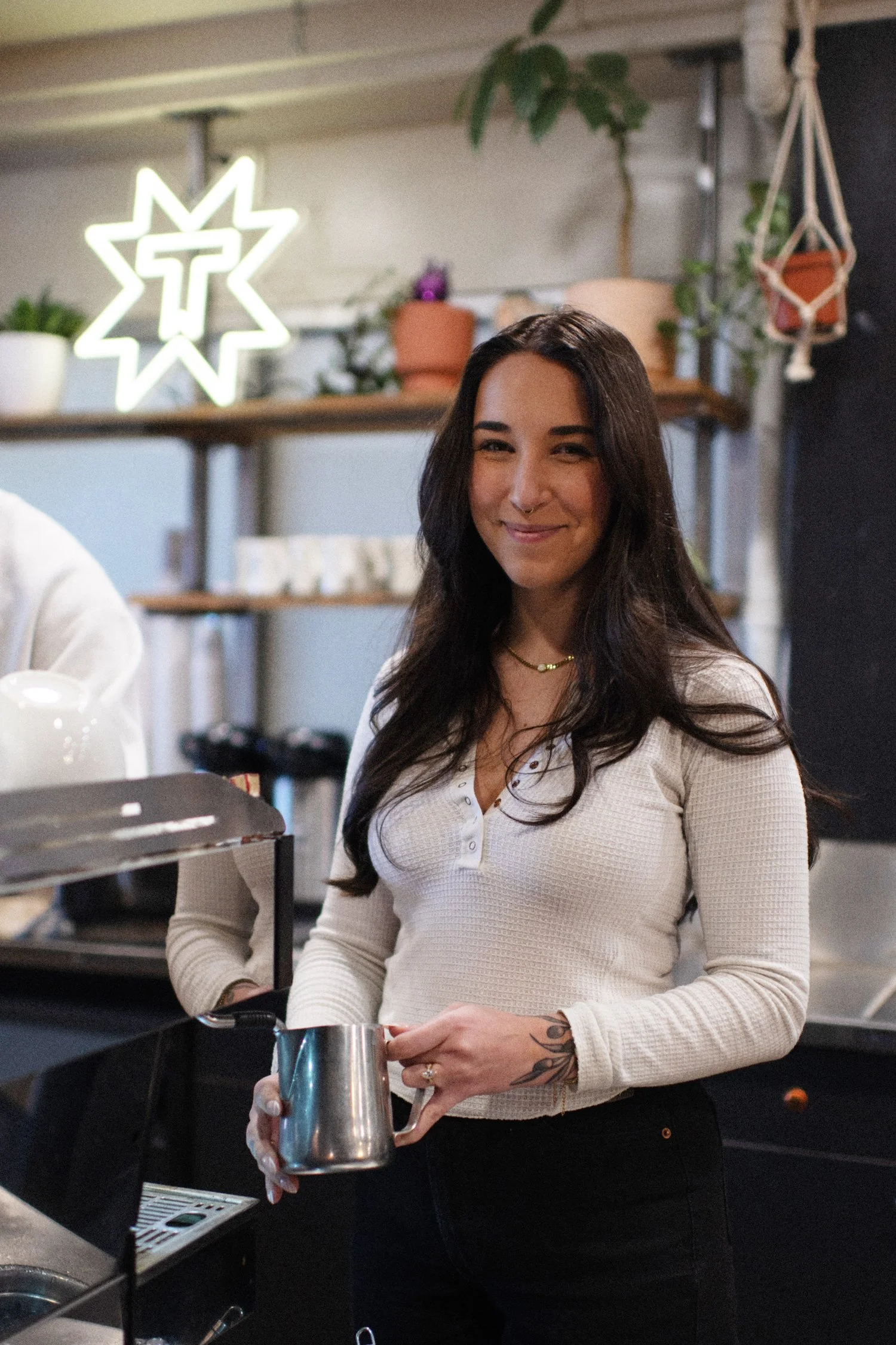 A woman with long dark hair and a nose ring, smiling, standing behind a coffee shop counter holding a metal pitcher. There are plants, a neon star-shaped sign with a cross, and shelves in the background.