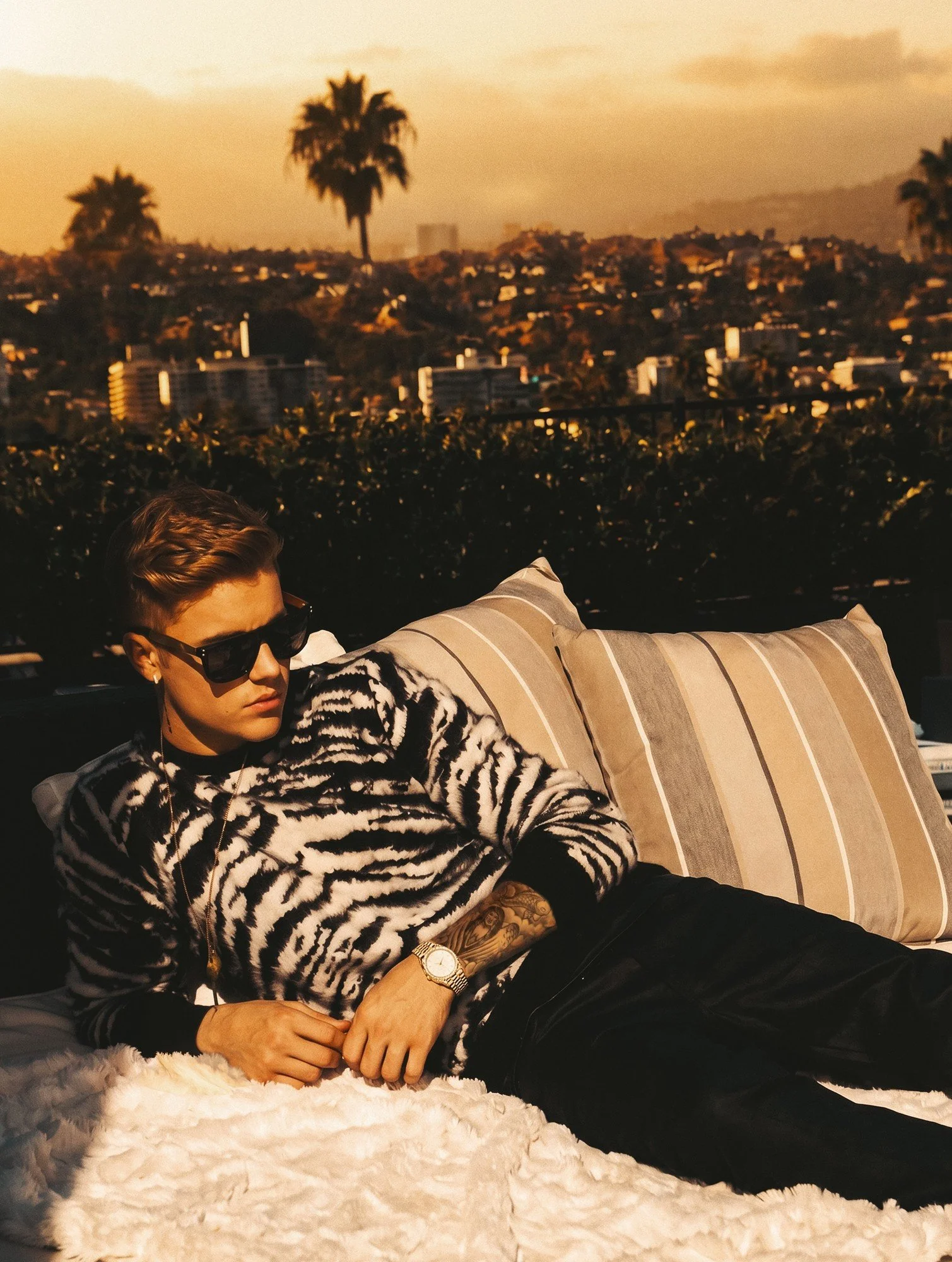 Justin Bieber by celebrity photographer Steven Gomillion. Wearing a black-and-white zebra-pattern sweater and dark sunglasses, he reclines on striped cushions and a white faux-fur throw, with palm trees and the Los Angeles skyline glowing behind him.