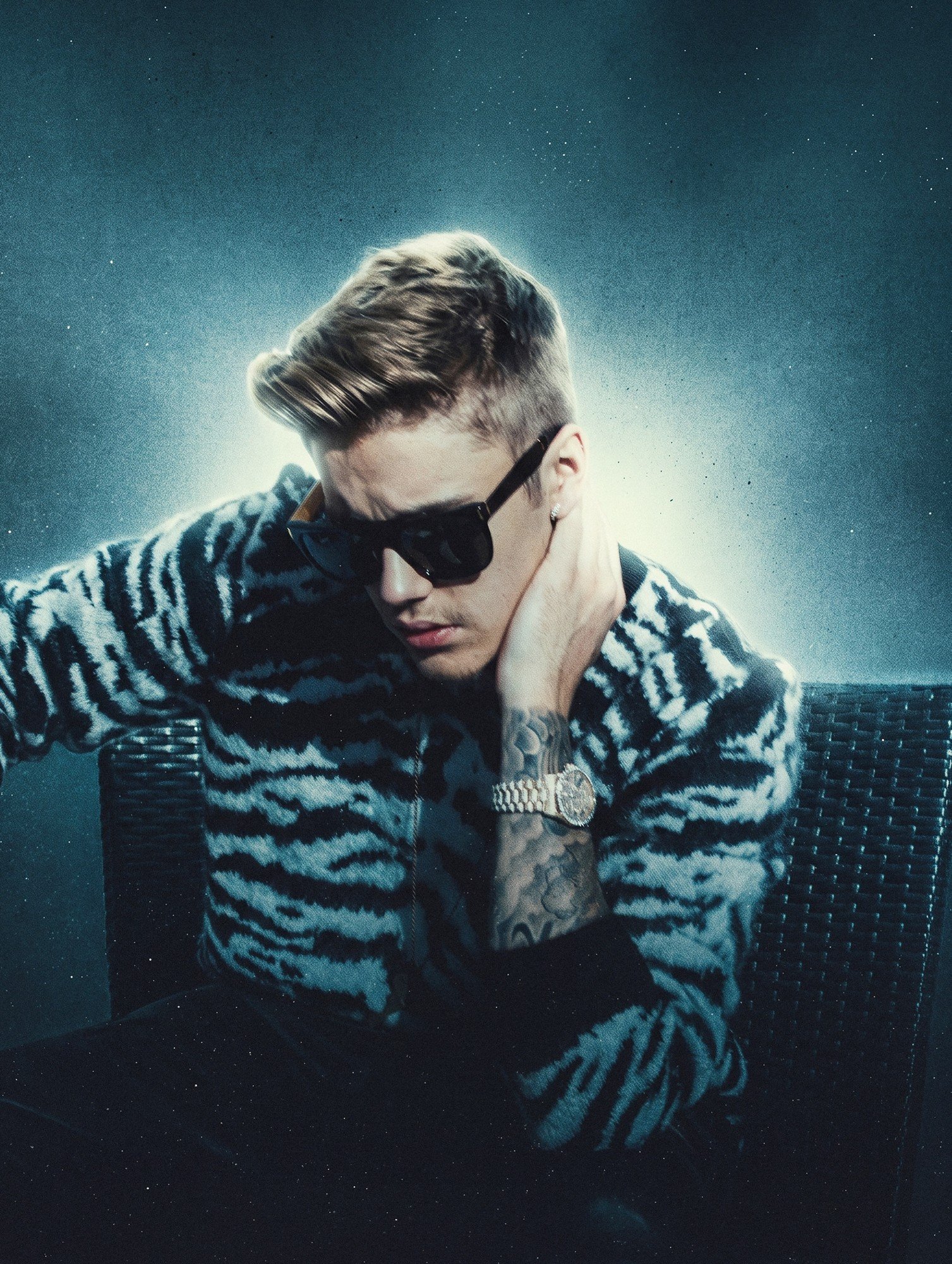 Justin Bieber by celebrity photographer Steven Gomillion. Wearing sunglasses and a zebra-pattern sweater, he sits in a wicker chair with one hand on his neck, a bright halo of backlight behind his head. Cool teal color grade with dust and grain textu