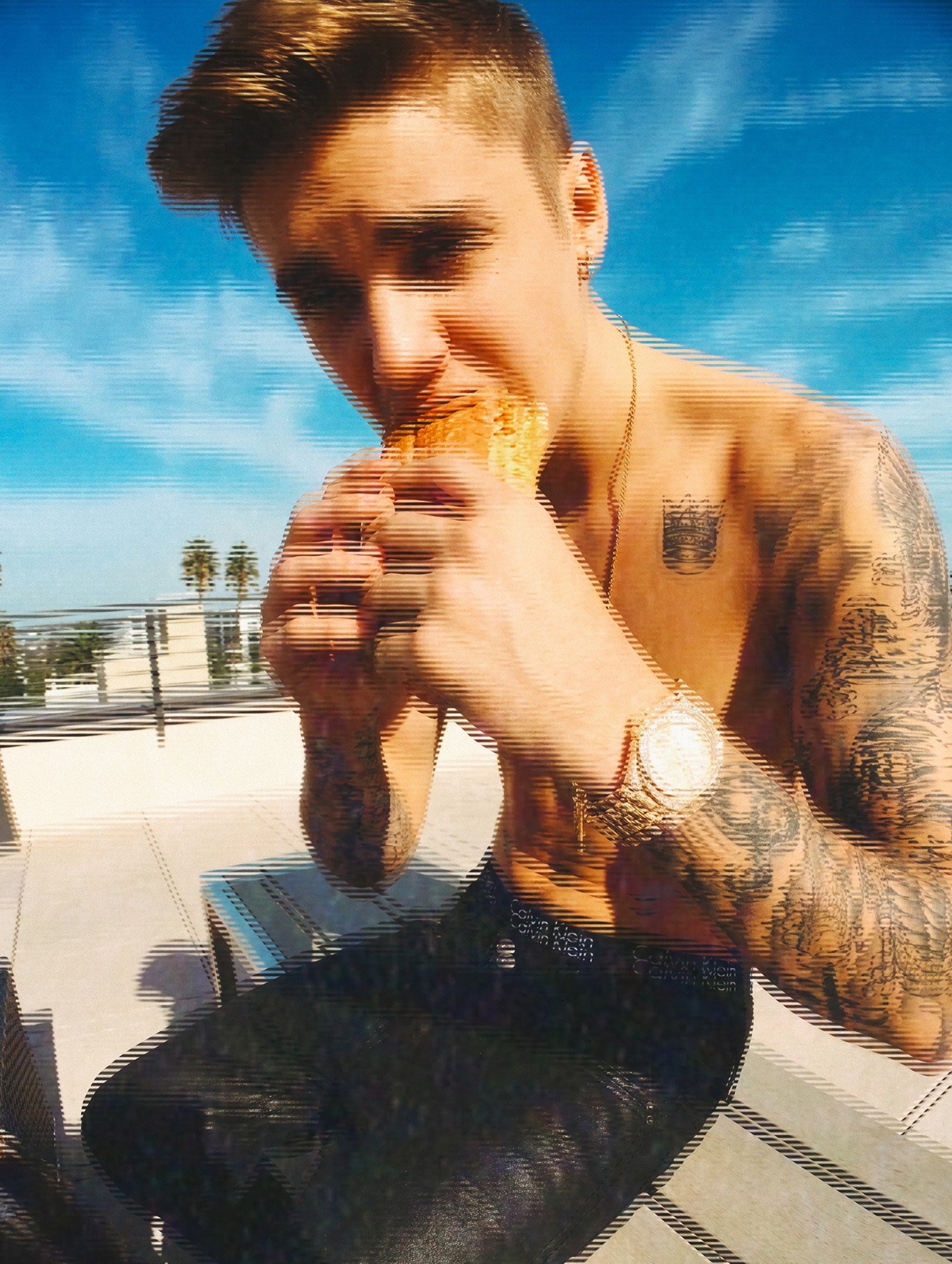 Justin Bieber by celebrity photographer Steven Gomillion. Shirtless with a Calvin Klein waistband, he bites into a sandwich close to the lens on a sunny rooftop, with palm trees and a bright blue sky behind. Saturated daylight color with a heavy hori