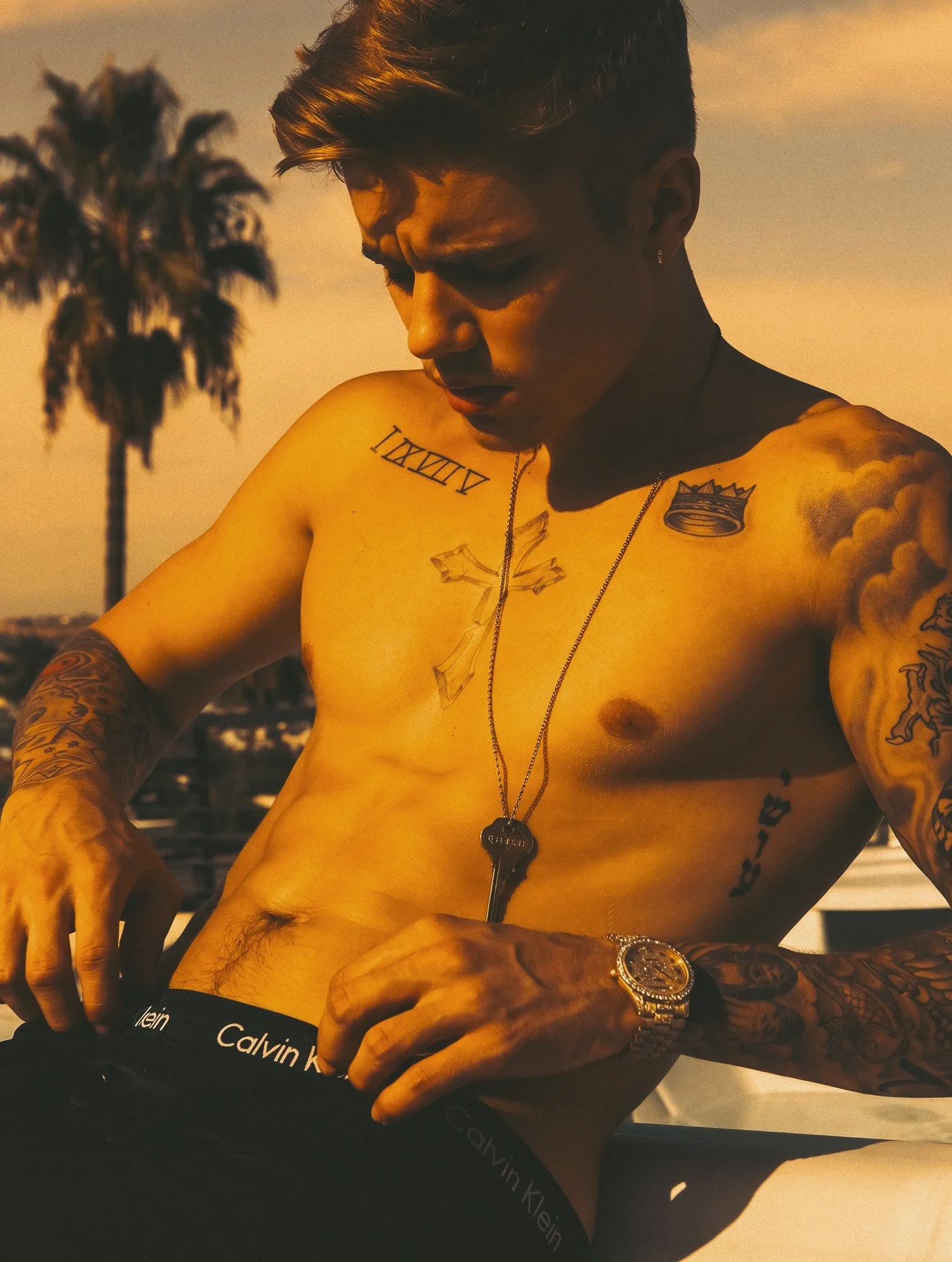 Justin Bieber by celebrity photographer Steven Gomillion. Shirtless, he adjusts a Calvin Klein waistband on a Beverly Hills rooftop, wearing a long key pendant and a diamond watch, with a palm tree and hazy sky behind him. Warm amber sunset color gra