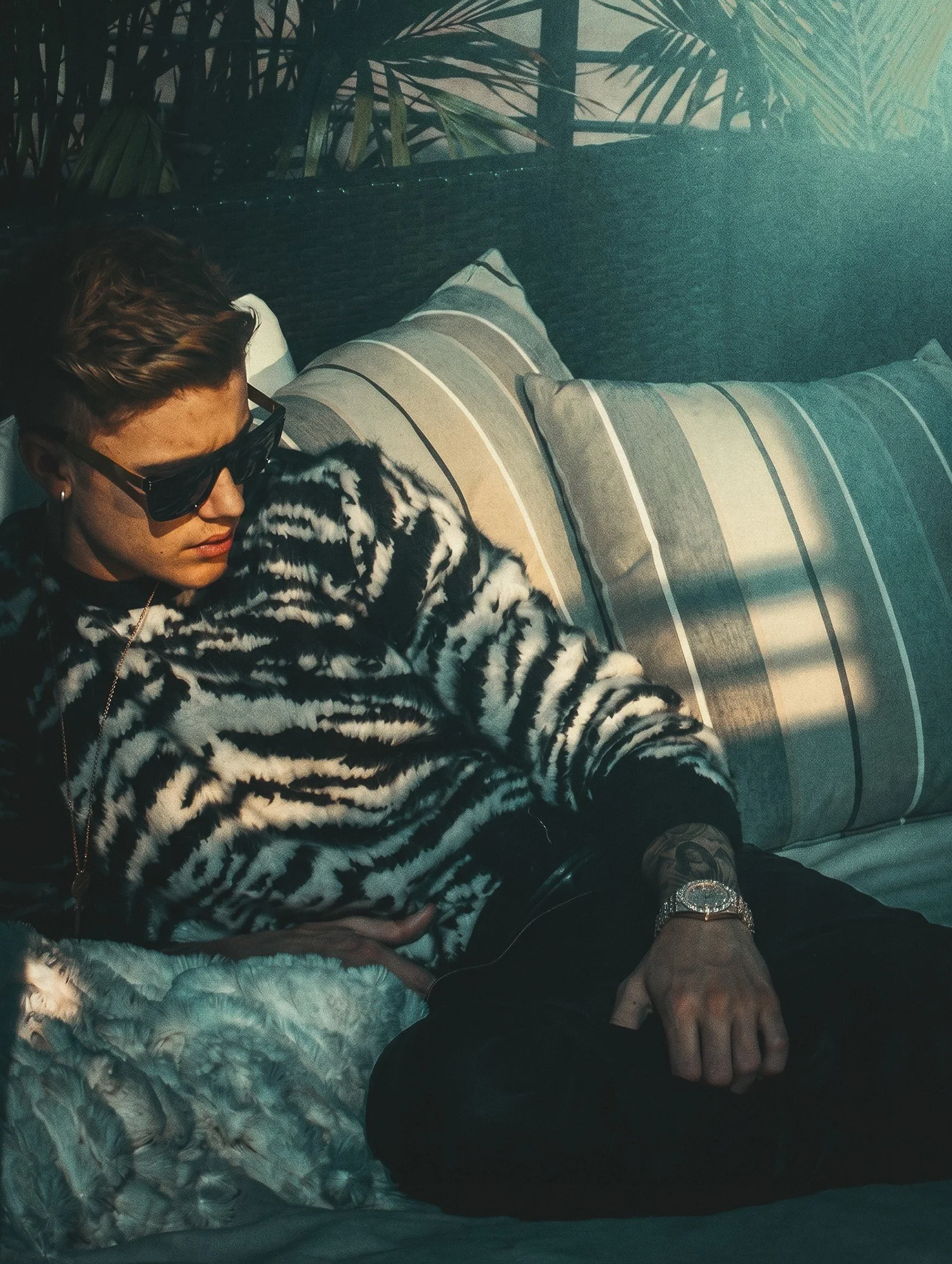 Justin Bieber by celebrity photographer Steven Gomillion. Wearing sunglasses and a zebra-pattern sweater, he leans back on striped outdoor cushions under palm fronds as hard, dappled sunlight falls across the frame. Cool, faded teal color grade.