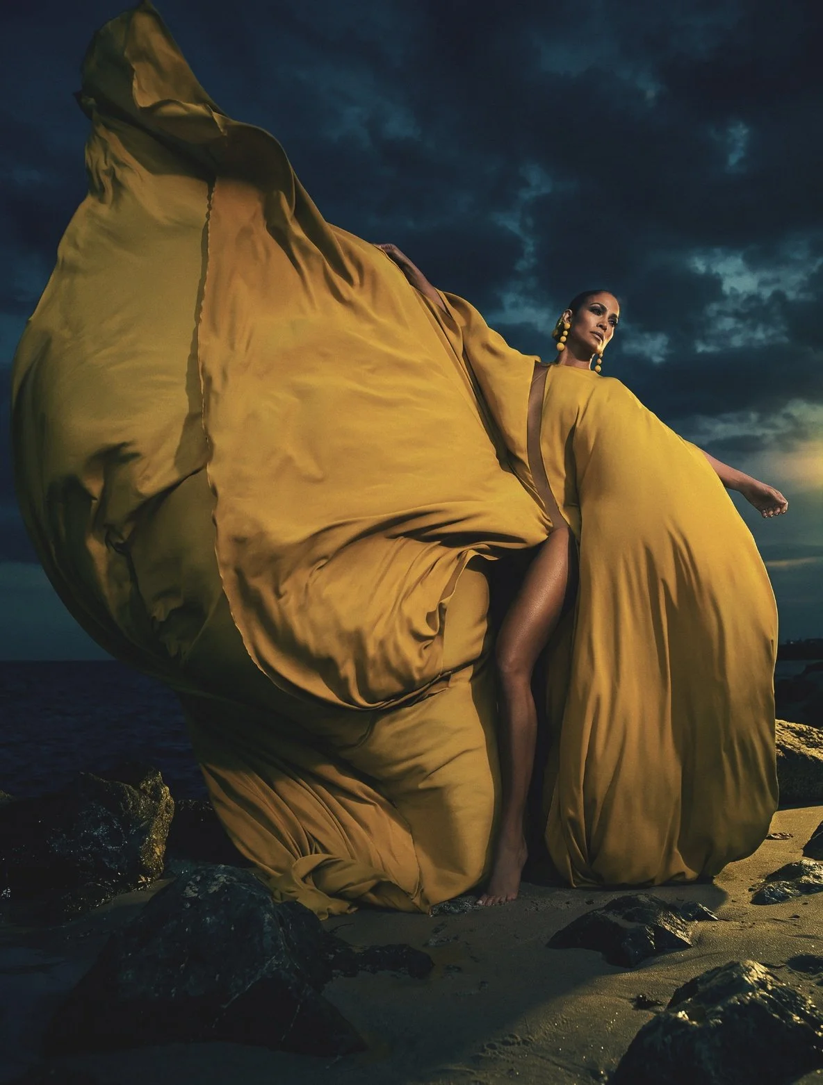 Jennifer Lopez in a billowing mustard-yellow gown on a rocky beach under a stormy sky by celebrity photographer Steven Gomillion