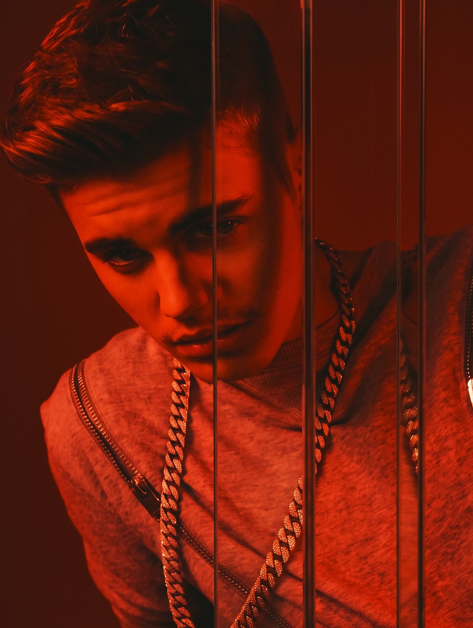 Justin Bieber by celebrity photographer Steven Gomillion. Close-up of him leaning toward the camera behind vertical glass panels that slice the frame, wearing a grey sweater and a chunky iced-out Cuban link chain. Hard, monochrome red color grade wit