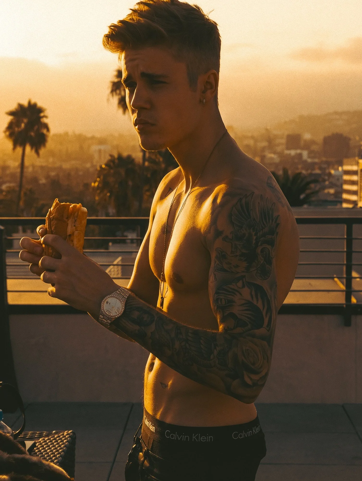 Justin Bieber by celebrity photographer Steven Gomillion. Shirtless and backlit on a rooftop terrace, he holds a sandwich, his tattooed arm and diamond watch catching the light, a Calvin Klein waistband visible, with palm trees and a hazy city skylin