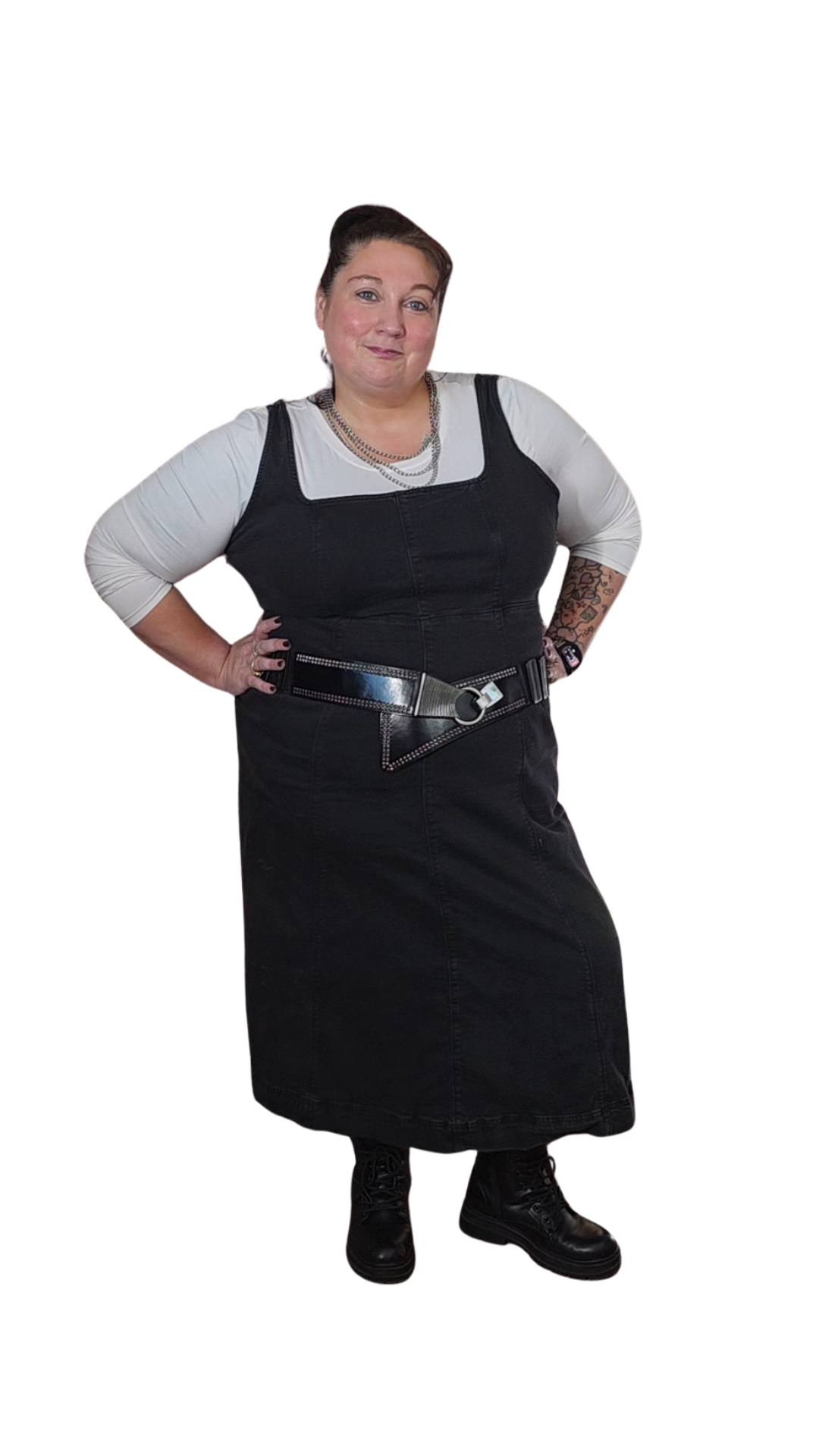 A woman wearing a black dress with a white long-sleeve shirt underneath, a thick black belt, and black boots, standing with her hands on her hips.