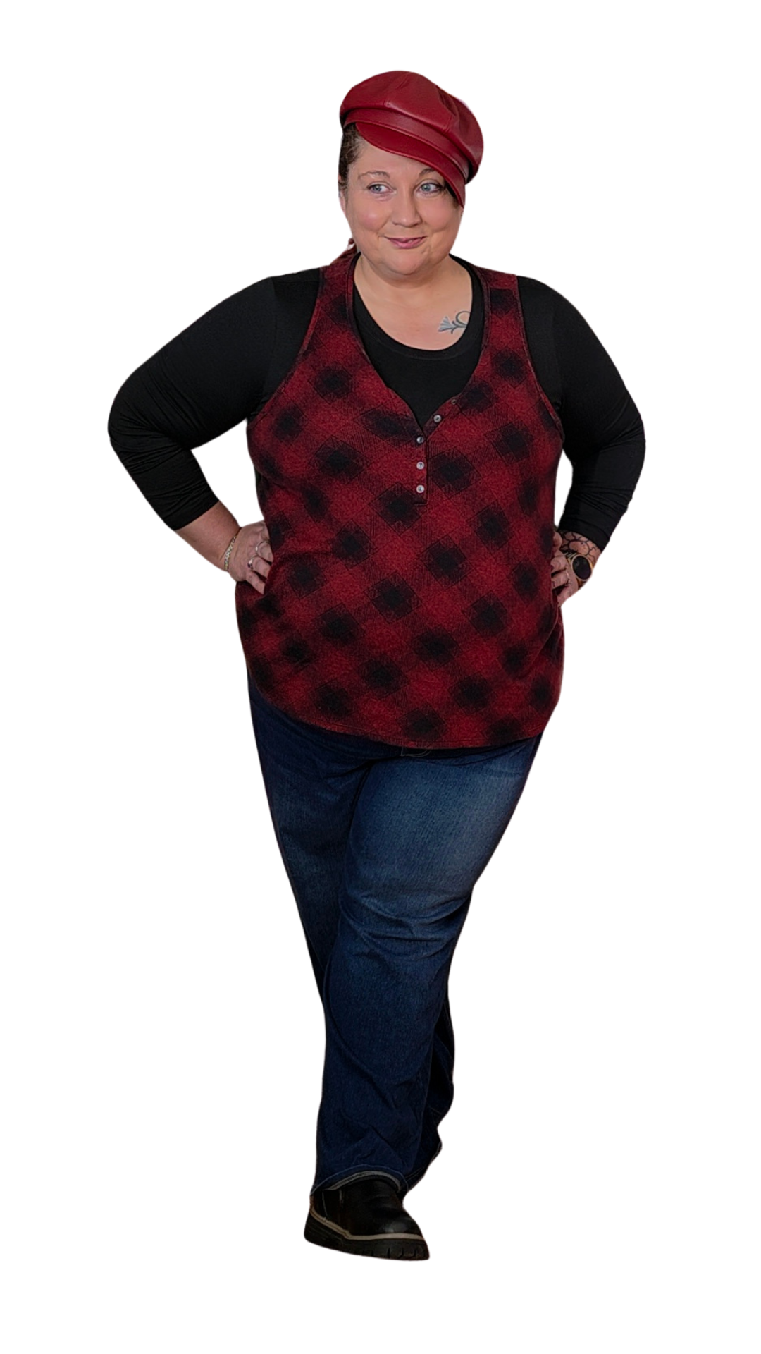 Woman wearing a red and black plaid sleeveless top, black long-sleeve shirt underneath, blue jeans, black shoes, and red leather cap, standing with hands on hips, smiling slightly.