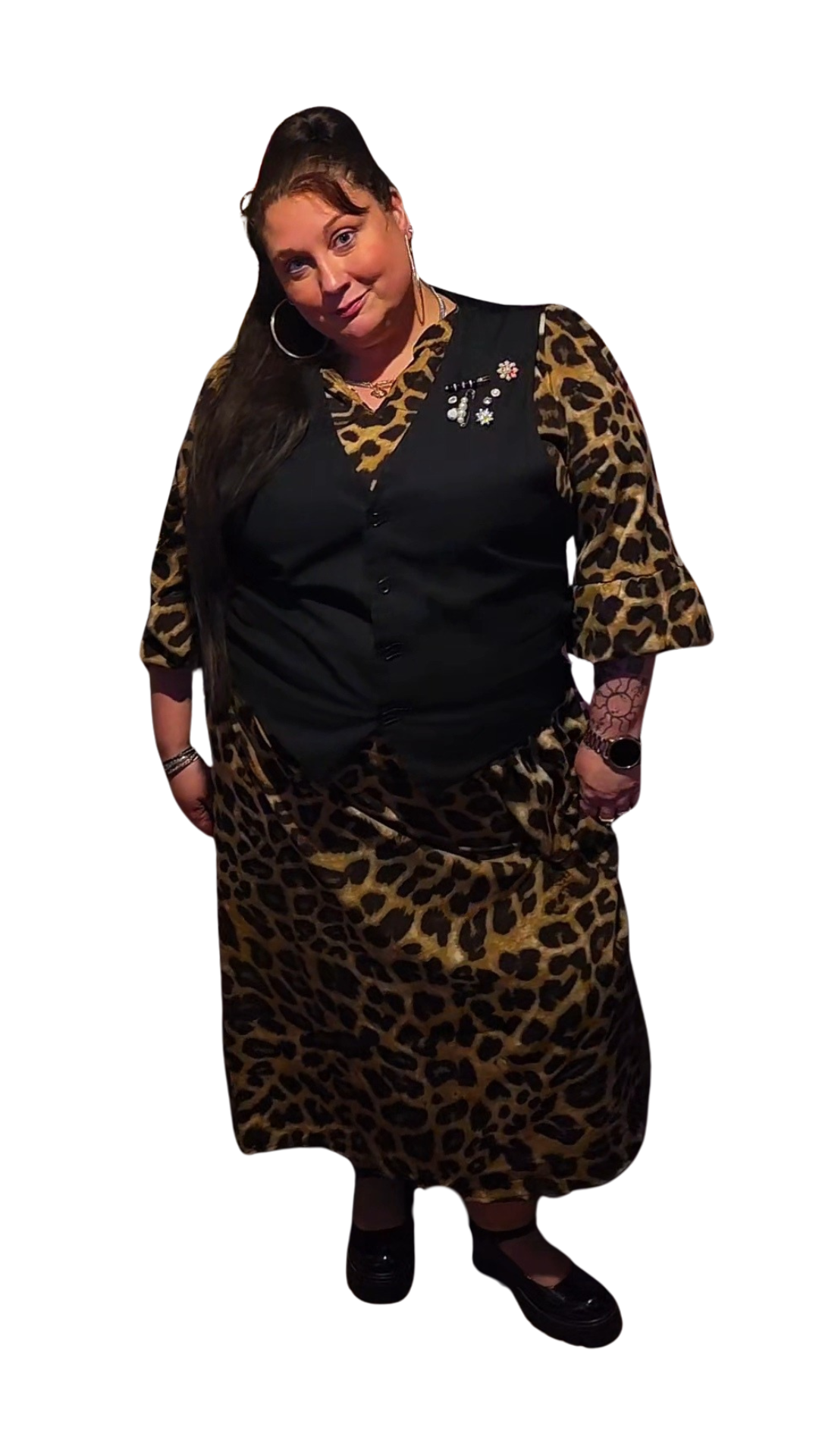 A woman in a leopard print skirt and a black vest with leopard print sleeves, wearing large hoop earrings, a watch, and jewelry, standing with her hands in her pockets against a plain white background.