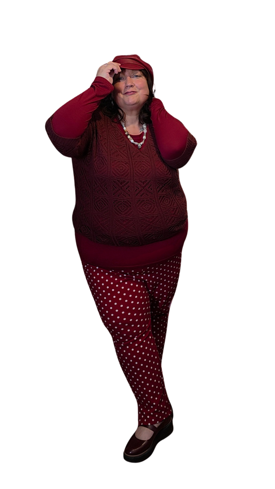 Woman in red and maroon clothing, wearing a red hat, patterned pants, and black shoes, posing with one hand on her hat.